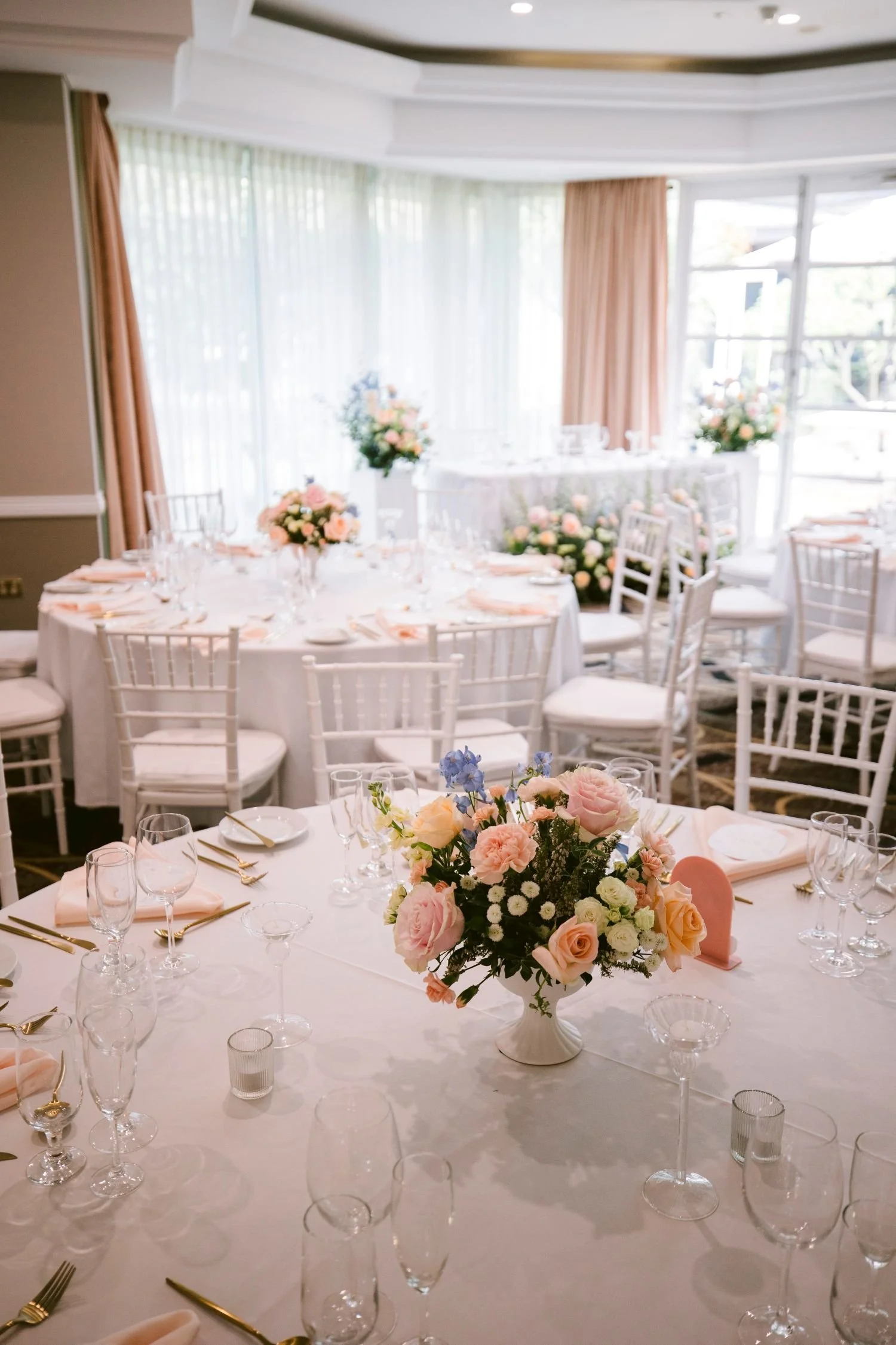 Elegant banquet table set for a celebration with a bouquet of pink and white flowers as the centerpiece, surrounded by white chairs. Well-lit room with large windows and sheer curtains.