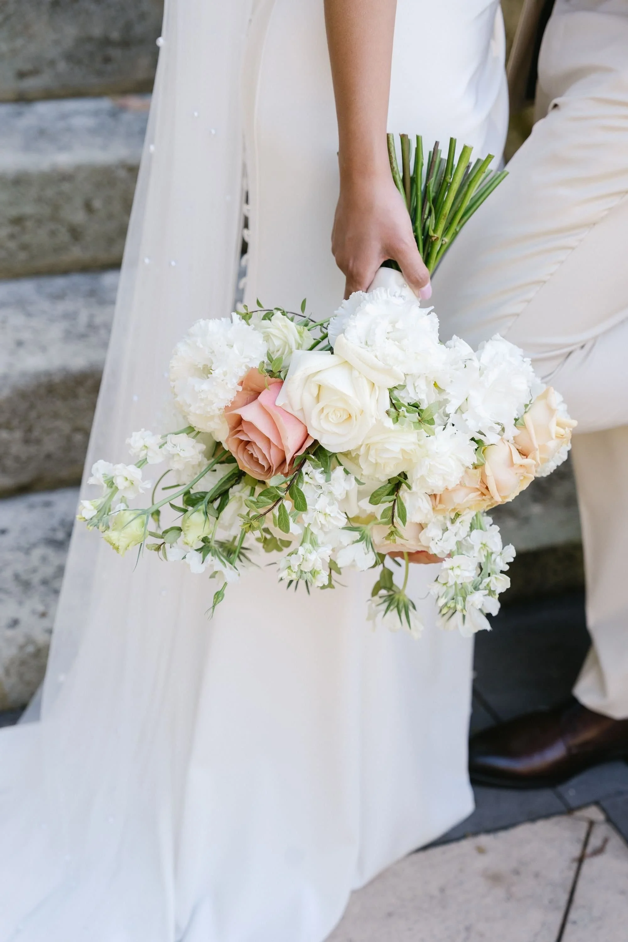 A bride wearing a white dress holding a bouquet of white, pale pink roses, and greenery. The person is standing near stone steps.