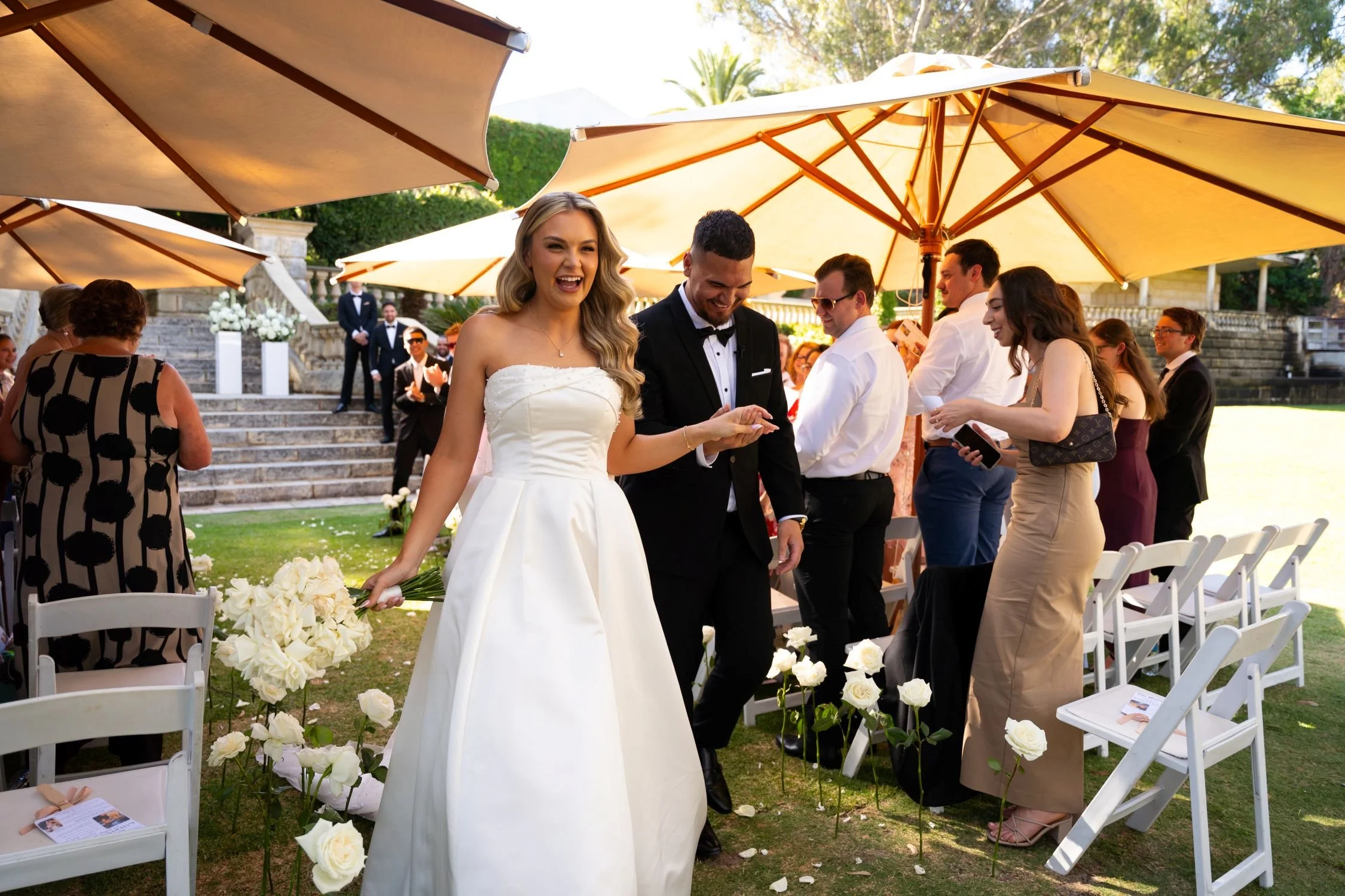 Wedding ceremony outside with a bride in a white gown and a groom in a tuxedo, surrounded by guests, under large yellow umbrellas on a grassy area with white chairs and floral decorations.