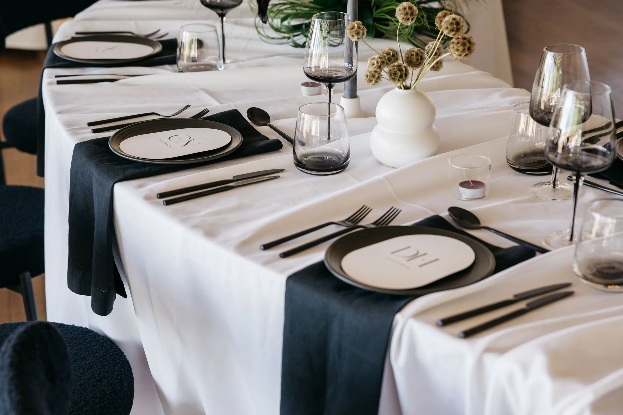 Elegant dining table set with black and white plates, black napkins, wine glasses, and a white vase with dried flowers.