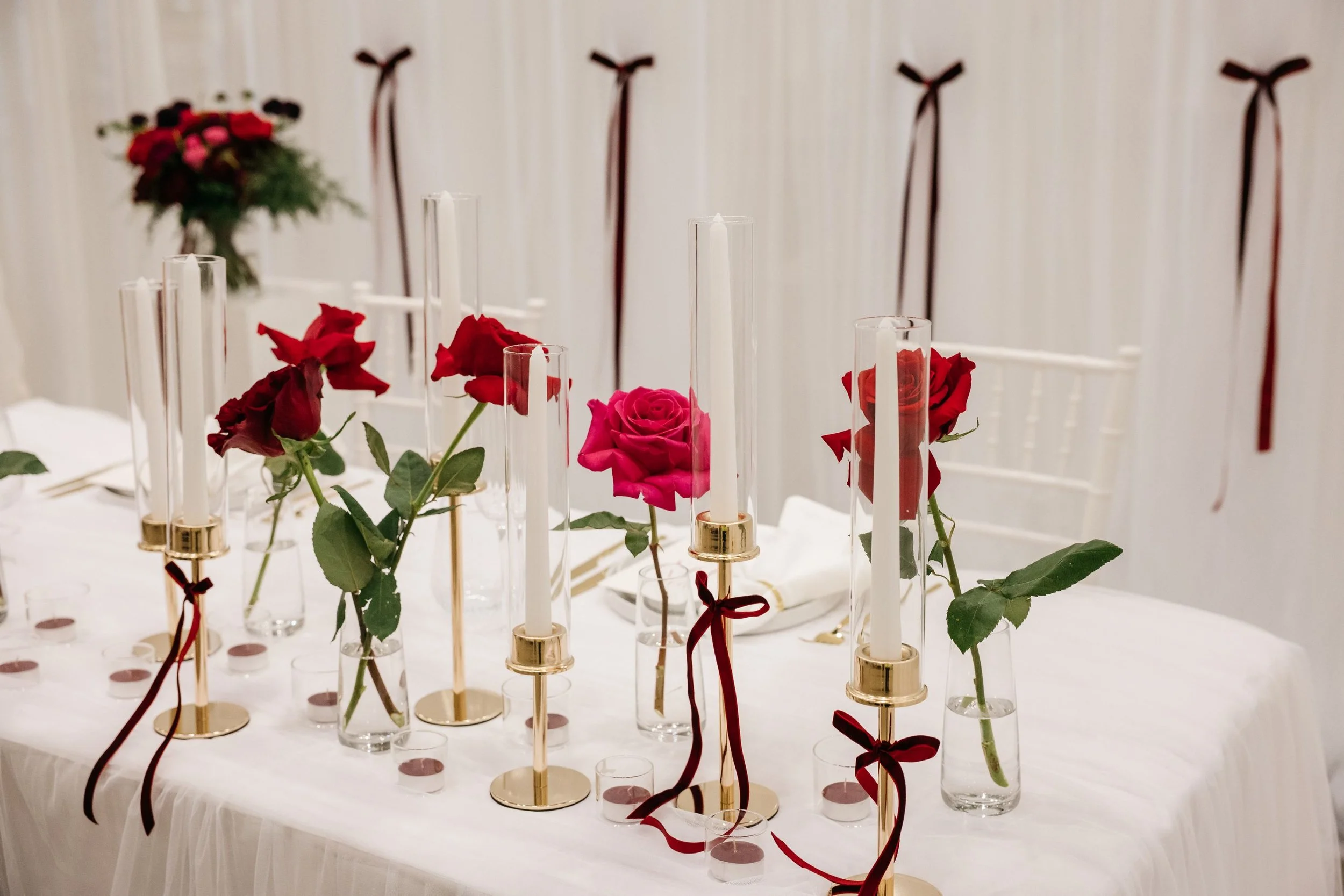 A sweetheart bridal table with red and pink roses in glass vases and tall white candles in gold holders, decorated with red ribbons, with a white curtain backdrop.