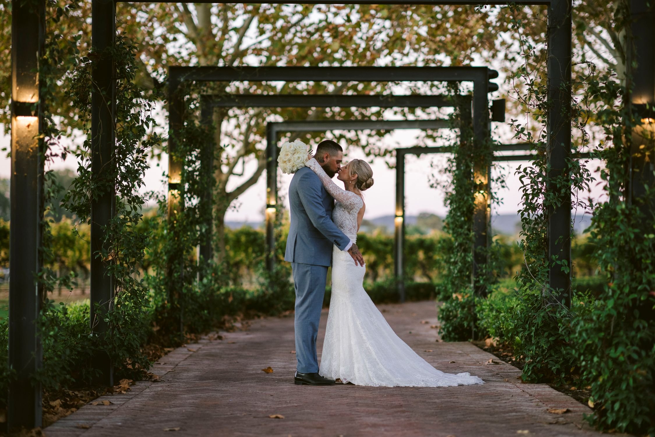 A bride and groom sharing a kiss under a trellis at sunset, trees with autumn leaves in the background, outdoor wedding setting.