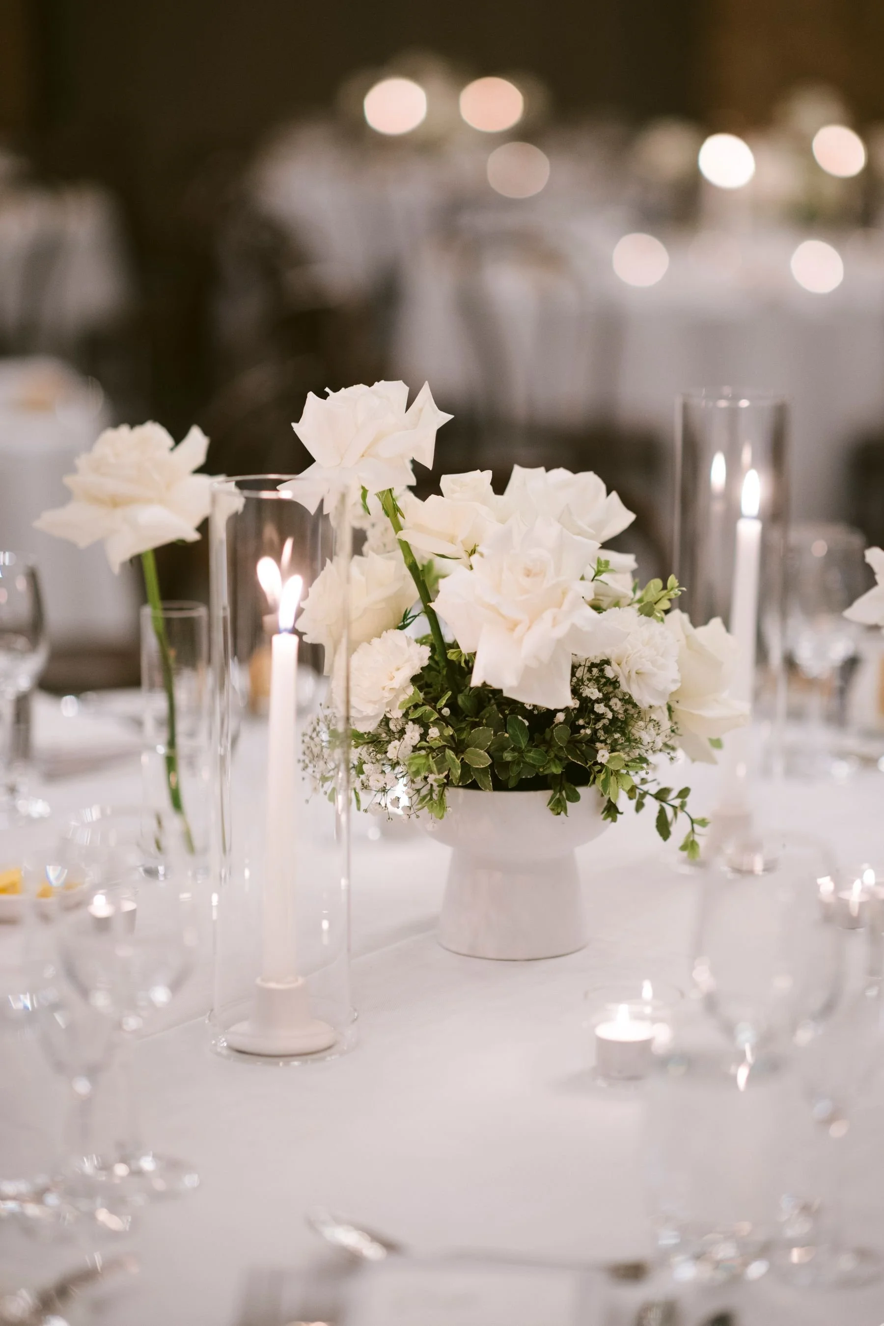 A floral centerpiece with white flowers in a white vase, surrounded by candles in glass holders on a table at a formal event.
