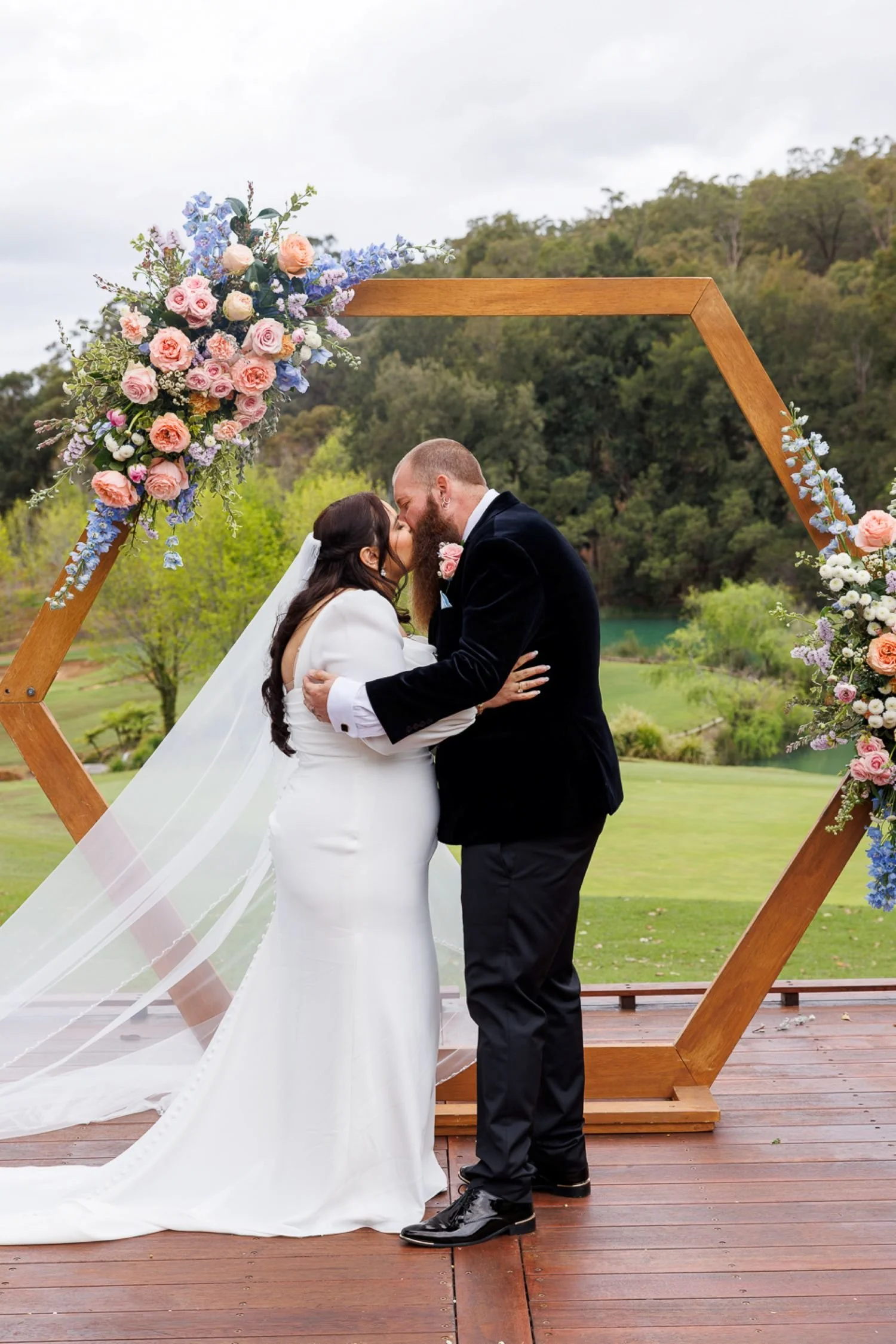 A bride and groom sharing a kiss under a wooden wedding arch decorated with pink, purple, and white flowers outdoors with a backdrop of trees and a lake.