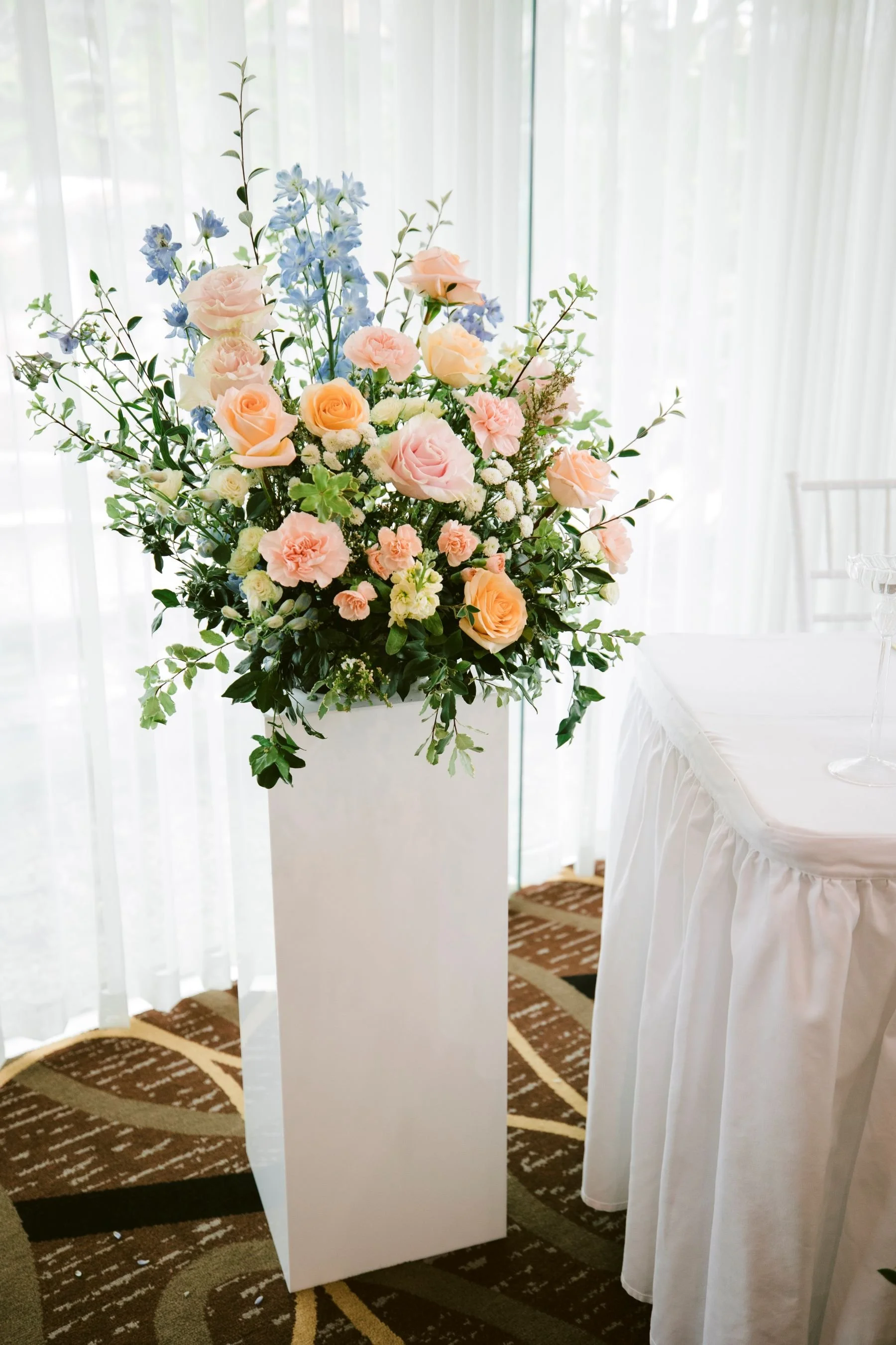 A large floral arrangement of pink roses, pale pink carnations, and blue delphiniums on a tall white stand in a room with sheer white curtains and a patterned carpet.
