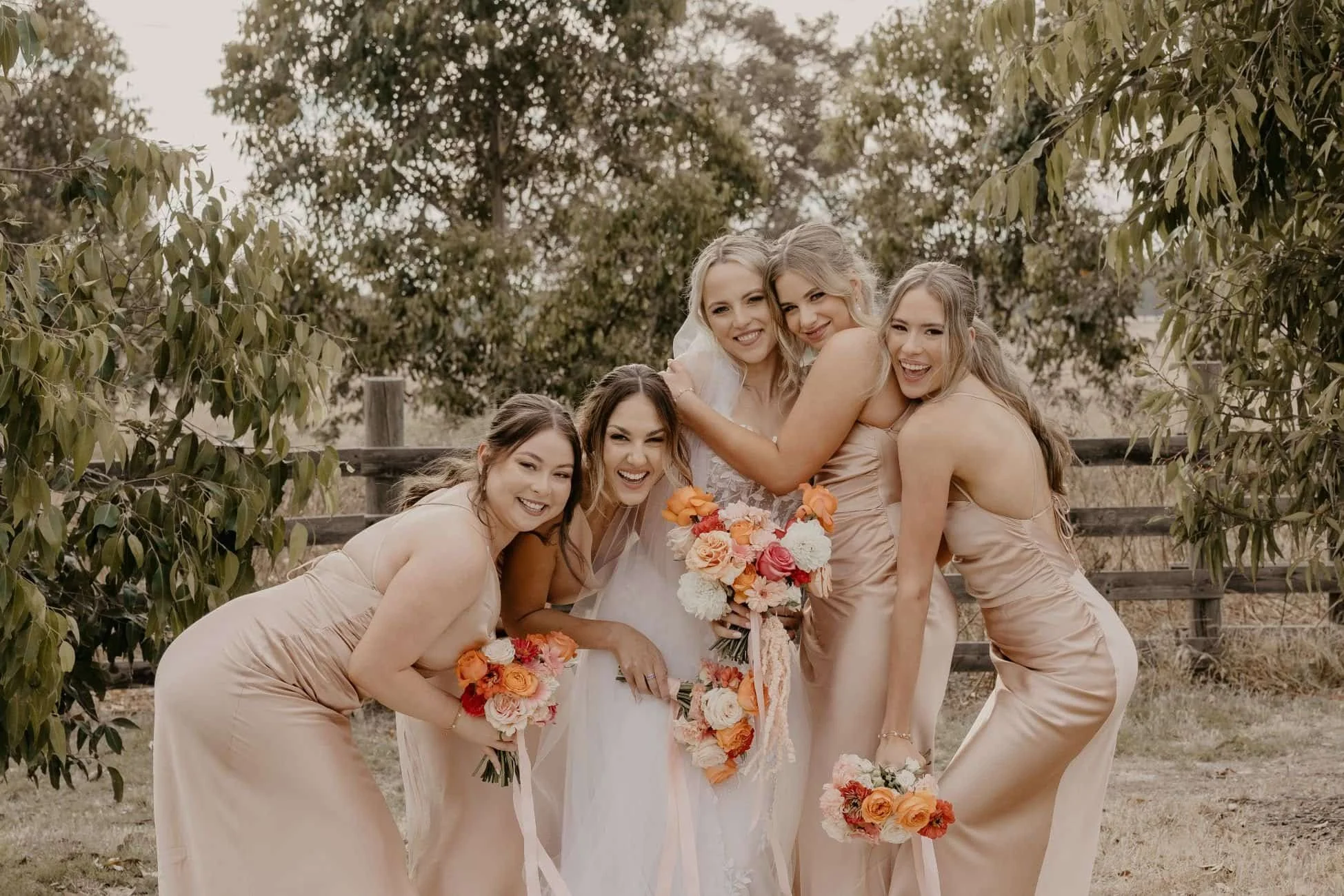 A bride and five bridesmaids smiling outdoors, dressed in matching nude-colored dresses, holding bouquets of flowers, with trees and a wooden fence in the background.