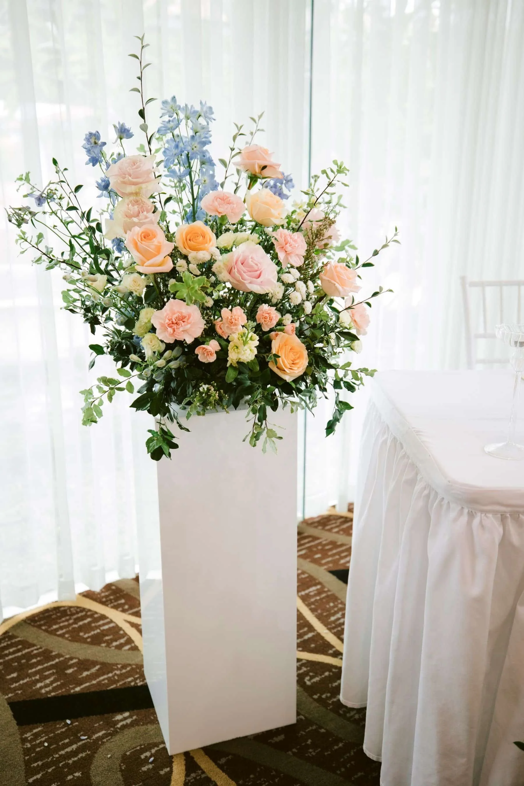 A large floral arrangement of pink, white, and blue flowers with green leaves stands on a white pedestal near sheer curtains, with a rounded table draped in white cloth and a chair in the background.