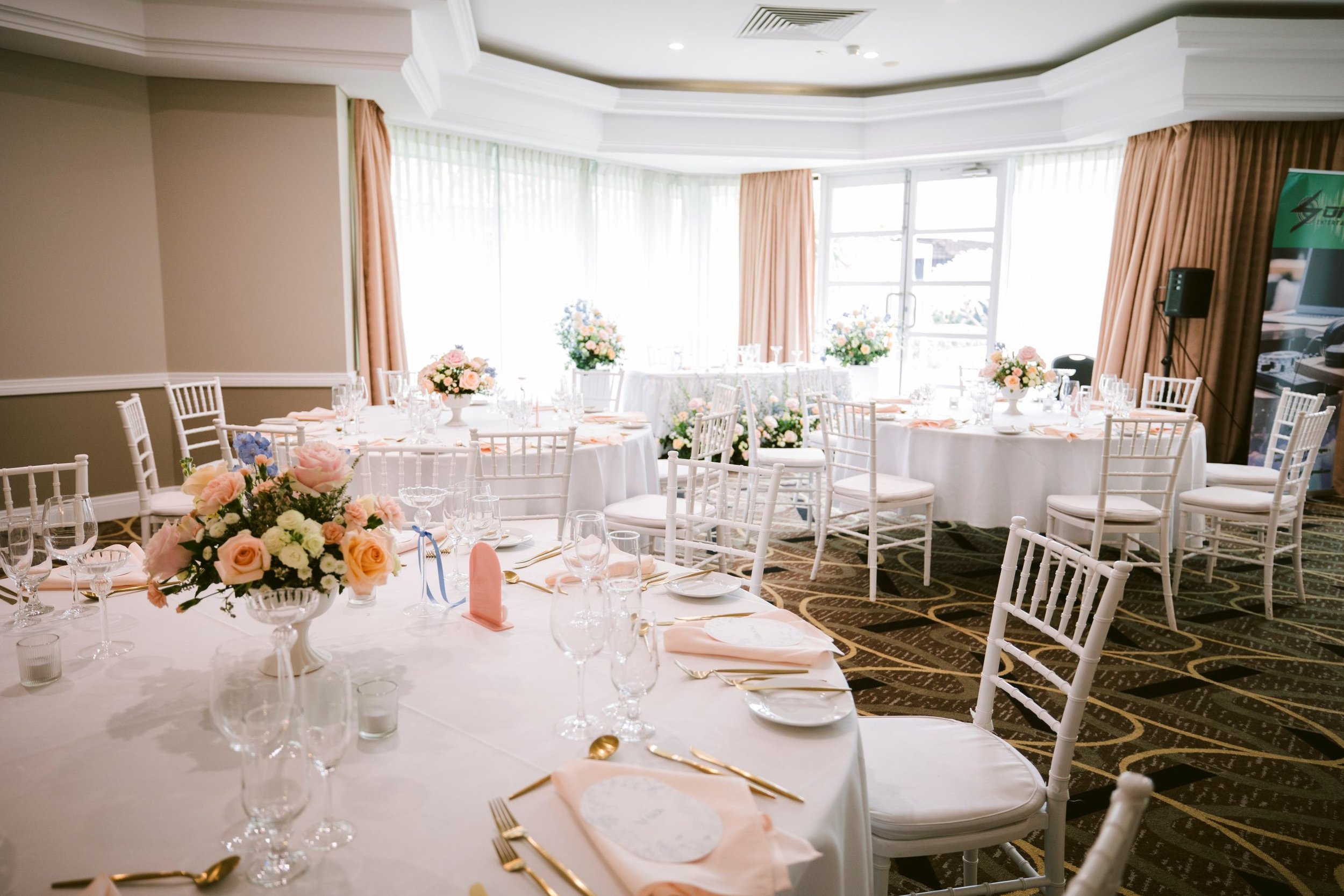Elegant banquet room setup with round tables covered in white tablecloths and decorated with pink and white flower centerpieces. The room features large windows with sheer curtains and pink drapes, white chairs around the tables, and table settings with glassware, gold utensils, and blush pink napkins. A speaker and a banner are visible to the right.
