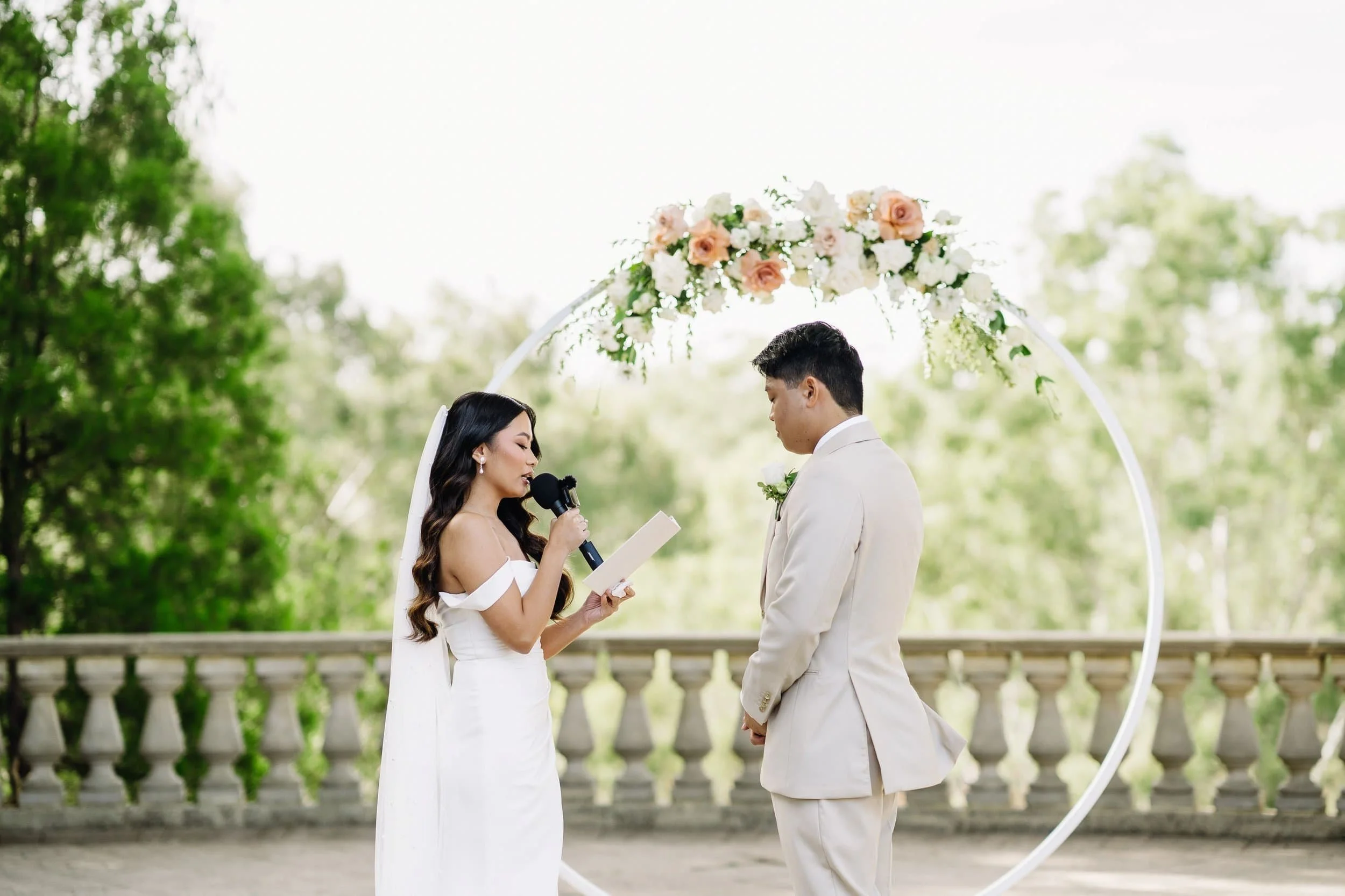 A couple getting married outdoors, with the bride reading vows and the groom listening.