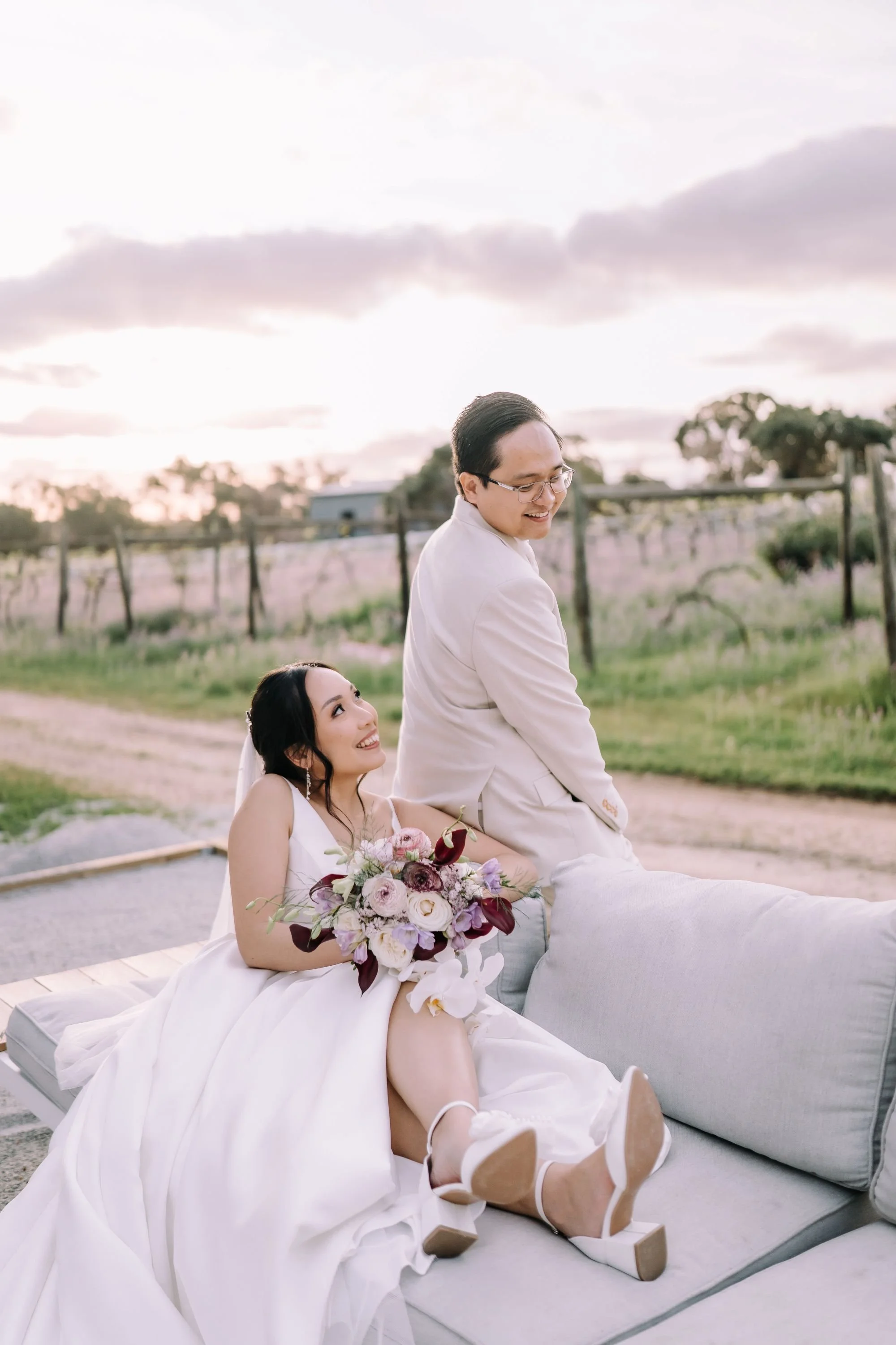 A bride and groom sitting outdoors on a cushioned bench, smiling and enjoying each other's company during their wedding day. The bride is holding a bouquet of flowers, and the groom is wearing glasses and a light-colored suit. The background features