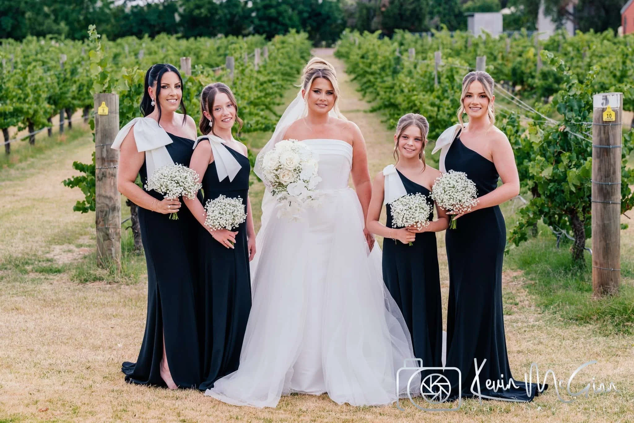 A bride and four young women, likely bridesmaids, standing in a vineyard. The bride is in a white wedding dress holding a bouquet, and the others are in matching black dresses with white bows, holding smaller bouquets.