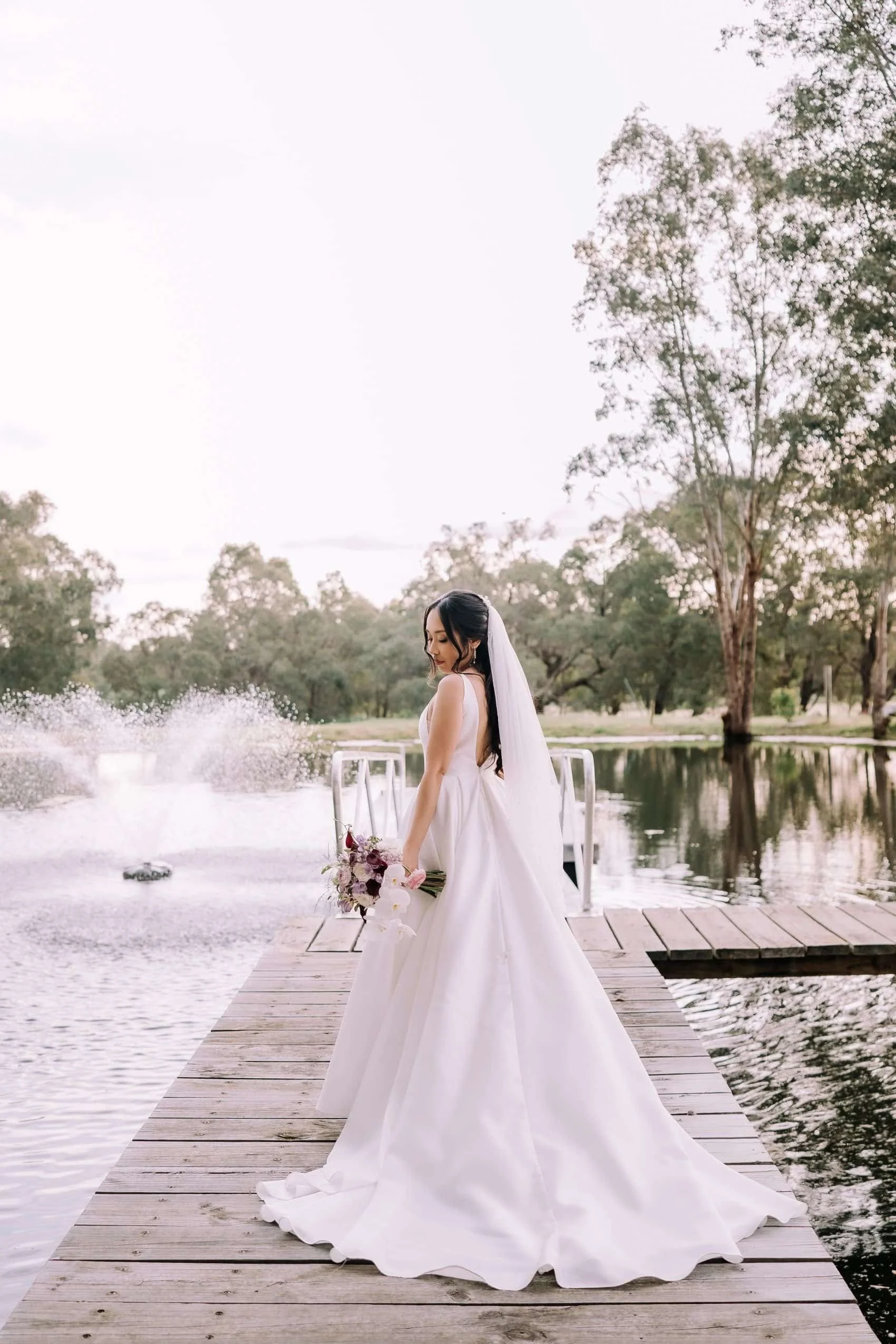 Bride in a white wedding gown standing on a wooden dock by a lake, holding a bouquet of flowers, with trees and a partly cloudy sky in the background.