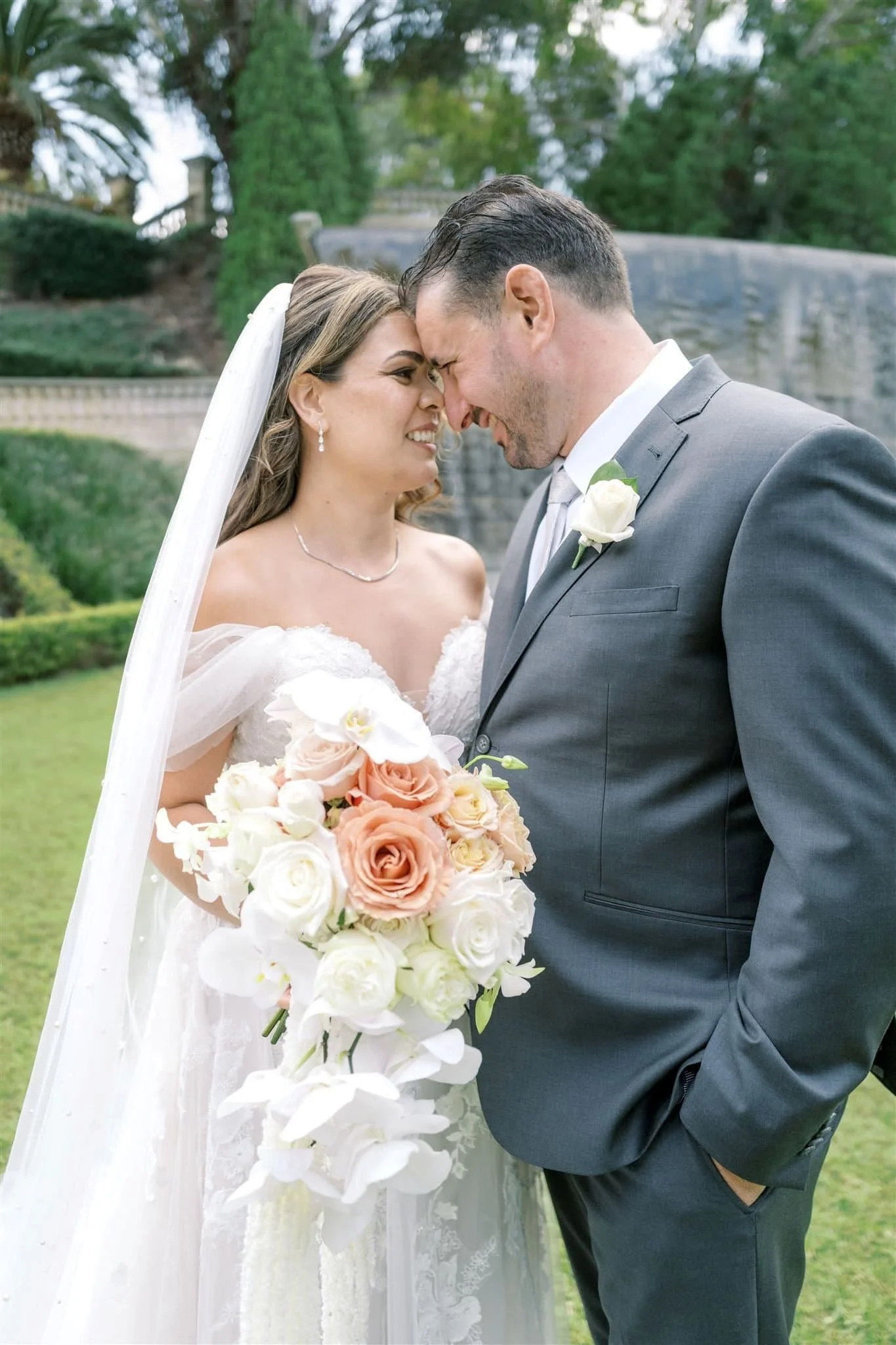A bride and groom on their wedding day standing close together with foreheads touching, smiling, outdoors in a garden with trees and a stone wall in the background. The bride is holding a large bouquet of white and blush roses.