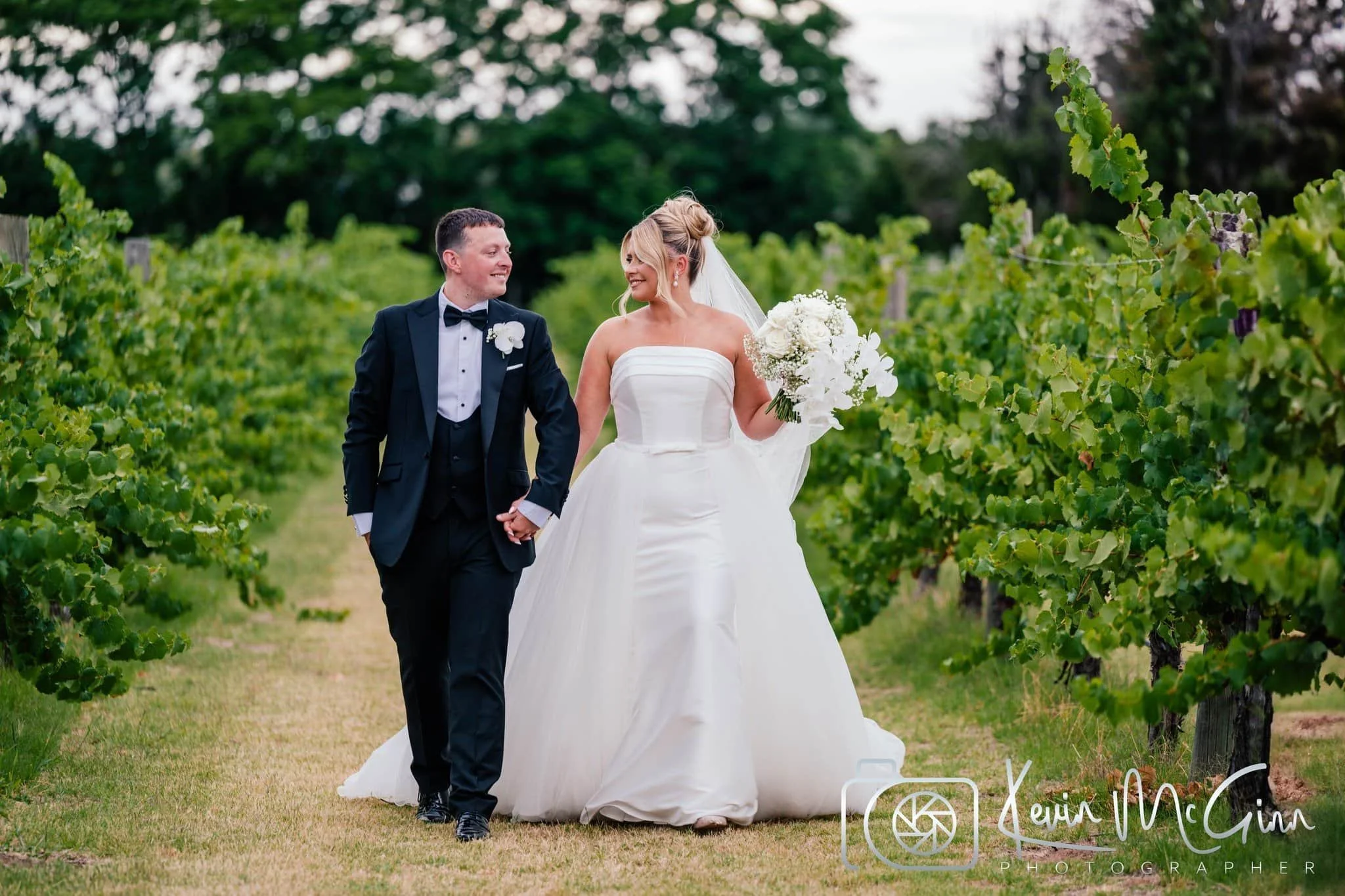 A bride and groom walking hand-in-hand through a vineyard, smiling at each other. The bride is in a white strapless wedding gown holding a bouquet, and the groom is in a black tuxedo with a bow tie. The vineyard is lush with grapevines and green leav