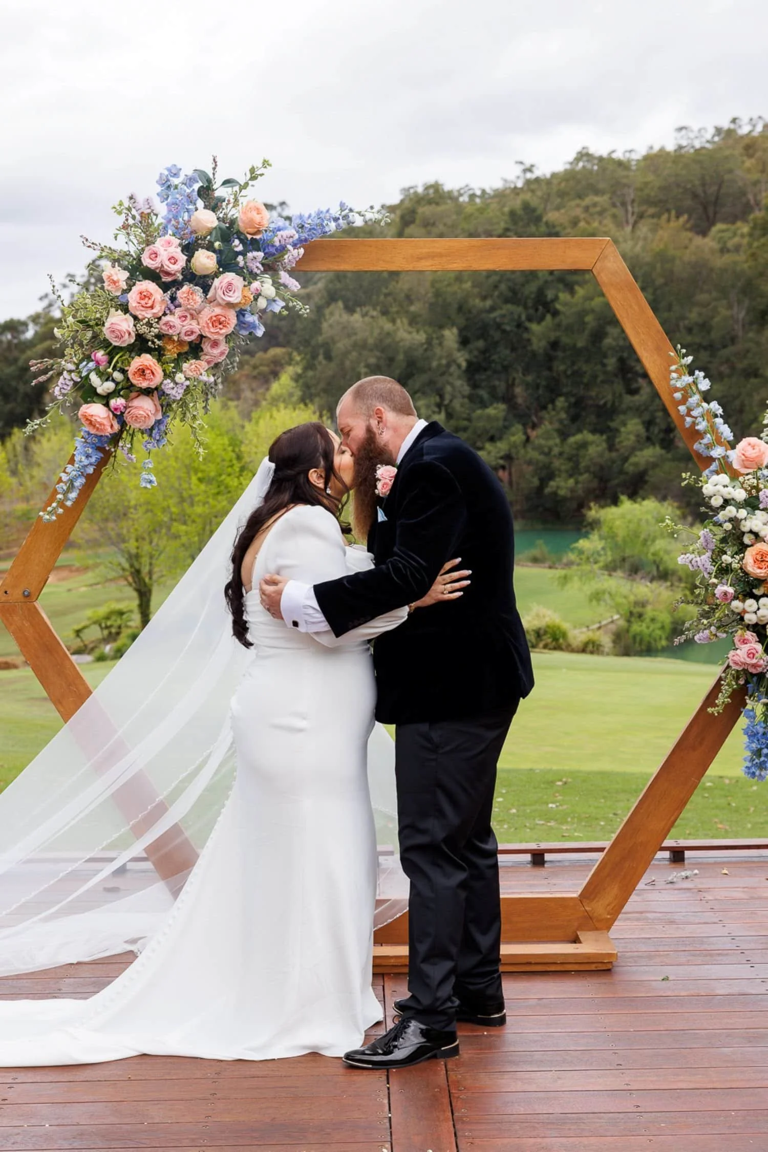 Couple dressed in wedding attire sharing a kiss at an outdoor wedding ceremony, decorated with a wooden hexagonal arch adorned with pink, peach, white, and purple flowers. The ceremony is set on a wooden platform with green landscape and trees in the