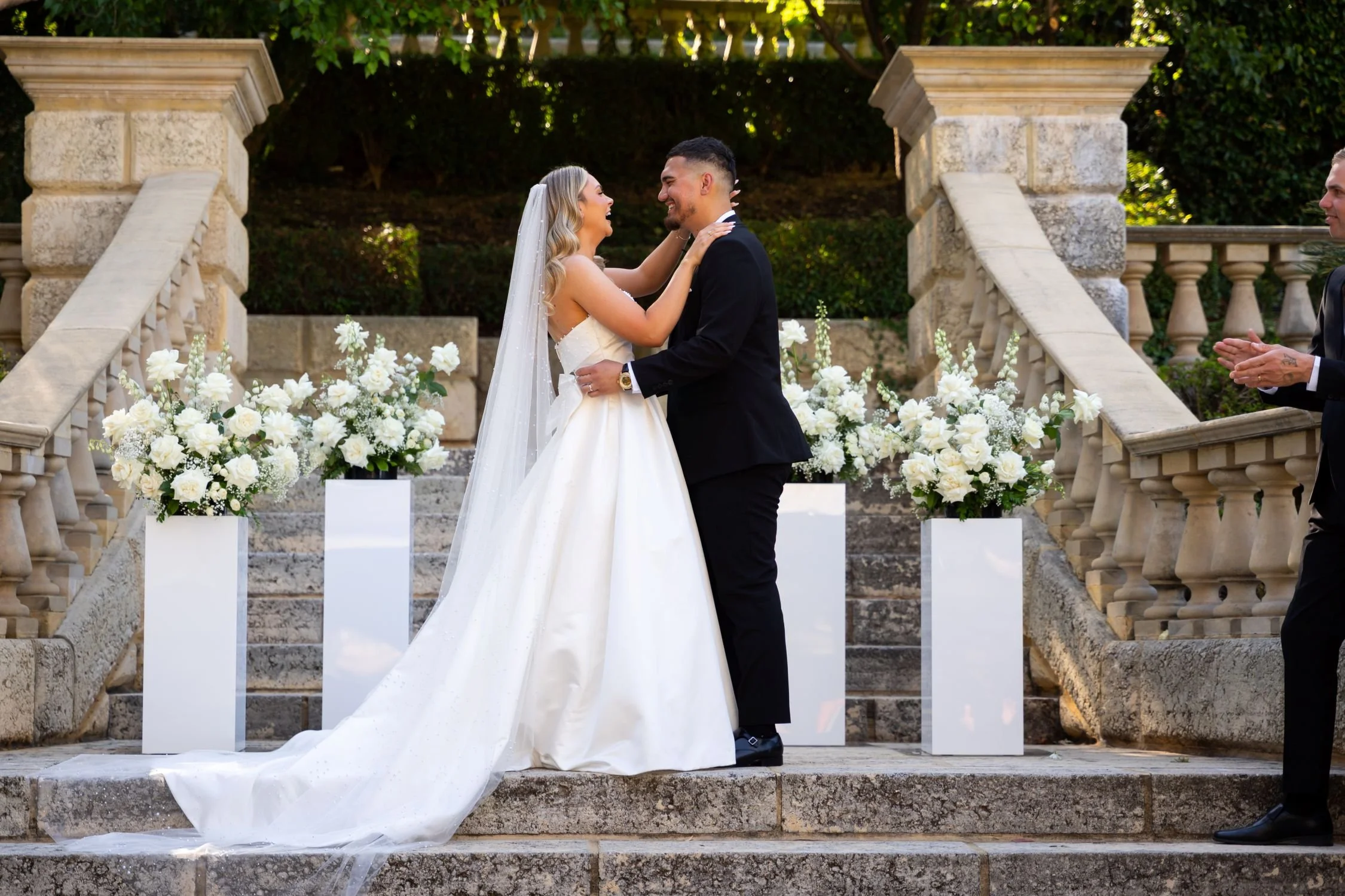 A bride and groom exchanging vows during their wedding ceremony outdoors on stone steps, surrounded by white floral arrangements and stone balustrades, with greenery in the background.