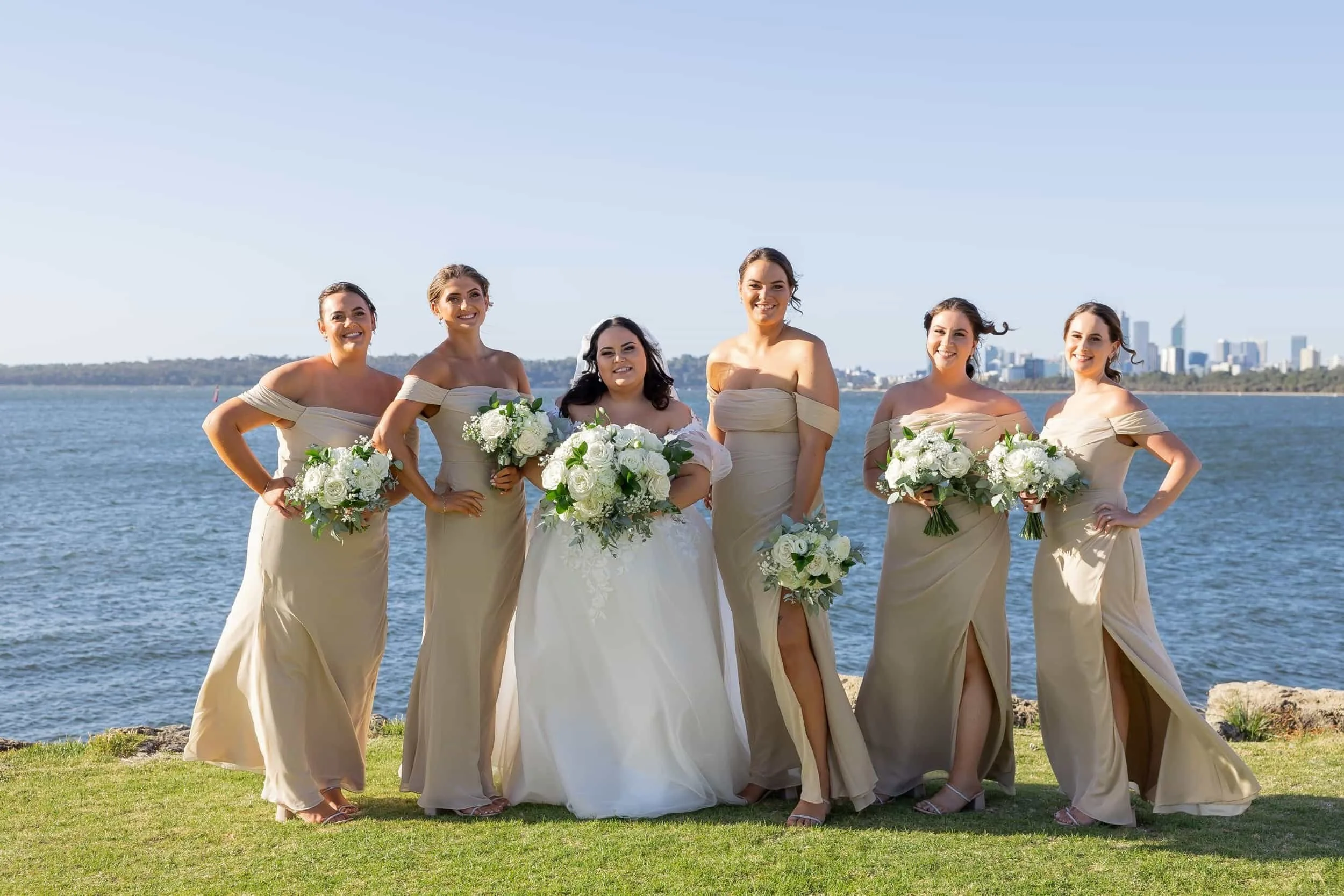 A bridal party of six women dressed in beige dresses, standing outdoors on grass near water with a city skyline in the background, smiling and holding white floral bouquets, appears to be at a wedding or celebration.