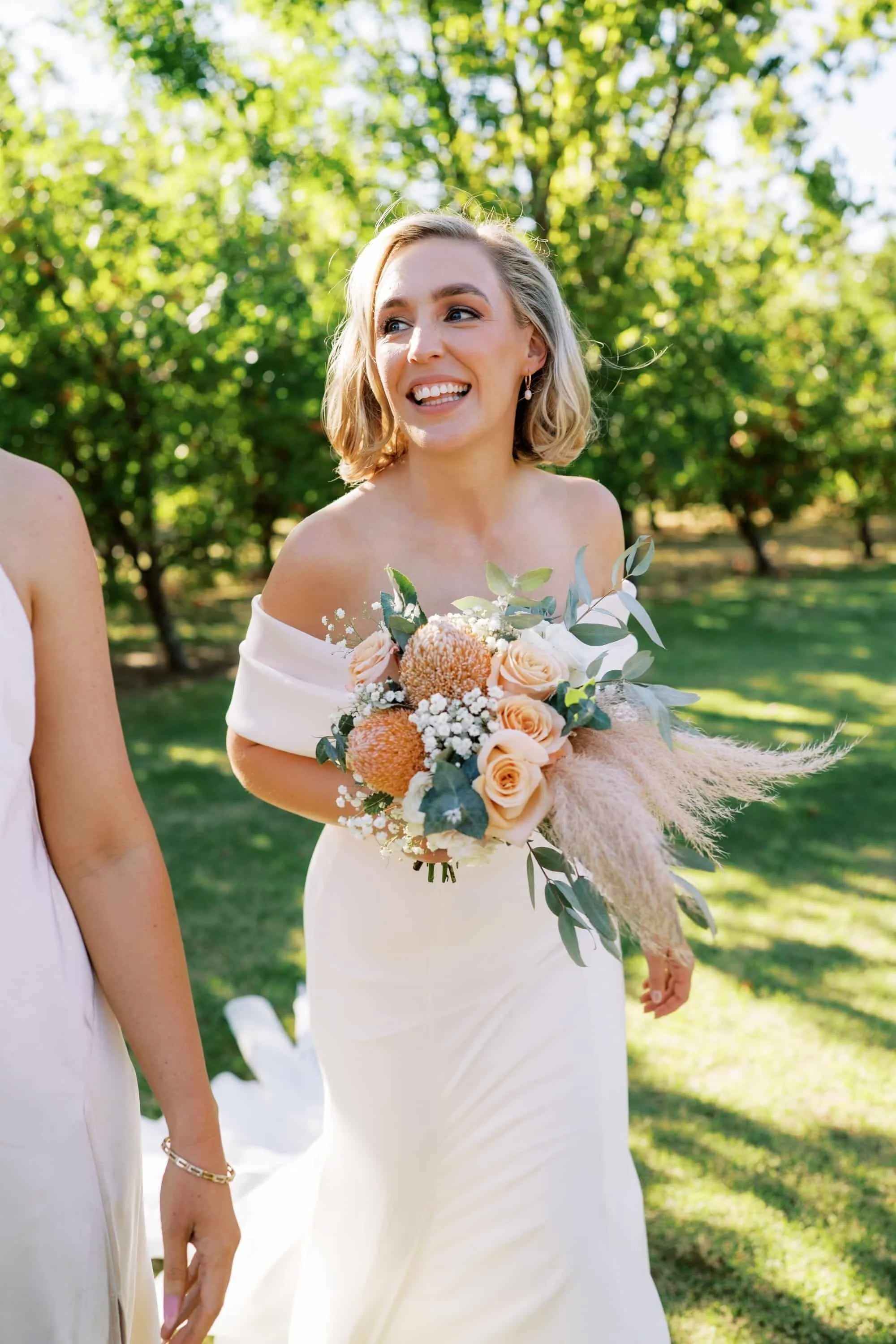 A woman in an off-shoulder white dress holding a bouquet of flowers, smiling outdoors with green trees in the background.