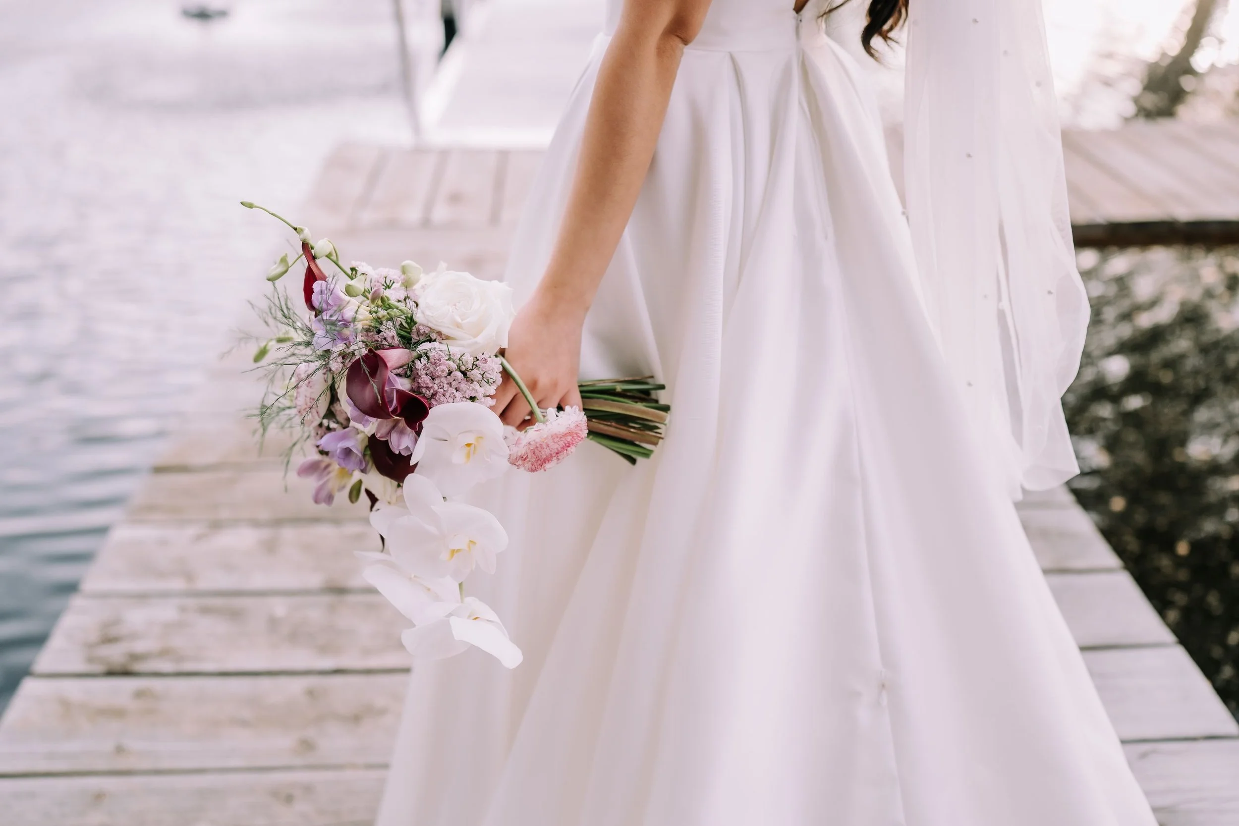A woman in a white dress holding a bouquet of flowers standing on a dock by a body of water.