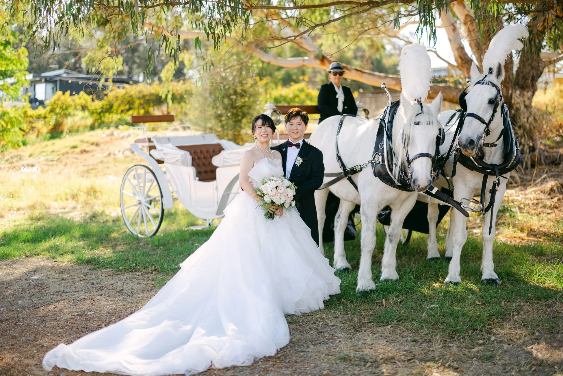 A bride and groom posing in front of a white horse-drawn carriage outdoors. The bride is wearing a white wedding gown and holding a bouquet, and the groom is dressed in a black tuxedo. A woman is sitting on the carriage in the background, and the set