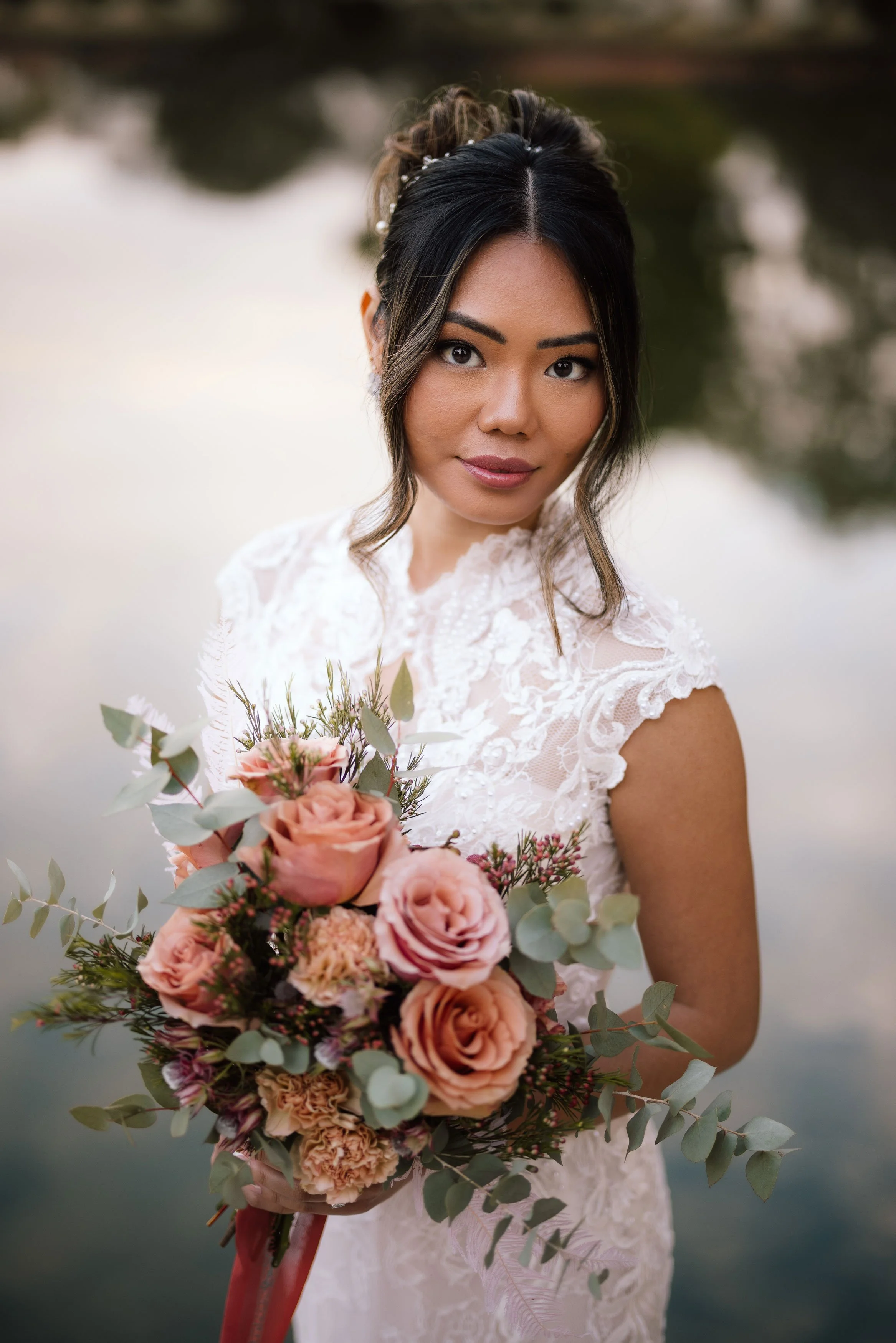 A woman in a white lace dress holding a large bouquet of pink and peach roses, eucalyptus leaves, and other flowers, standing outdoors near a body of water.
