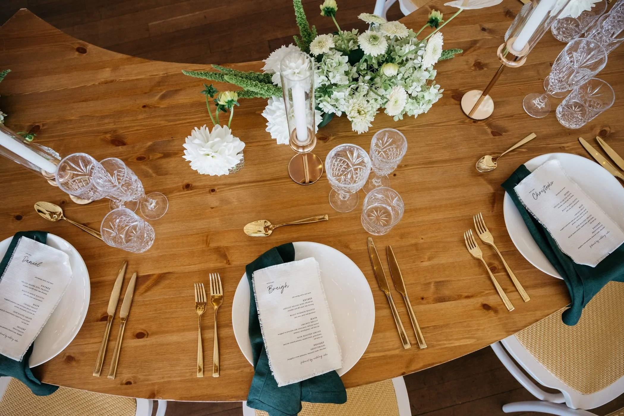A wooden dining table set for a formal meal, featuring white plates with folded napkins and gold utensils, crystal glasses, and a floral centerpiece with white flowers and greenery.