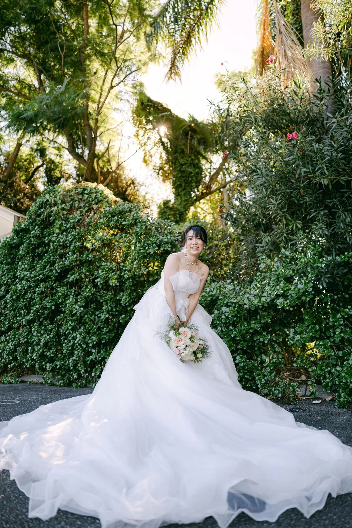 A bride in a white wedding gown holding a bouquet of flowers, standing outdoors among lush green bushes and trees with sunlight filtering through.