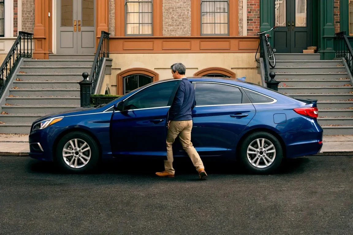 Man walking to a blue sedan parked on a city street with brownstone buildings in the background.
