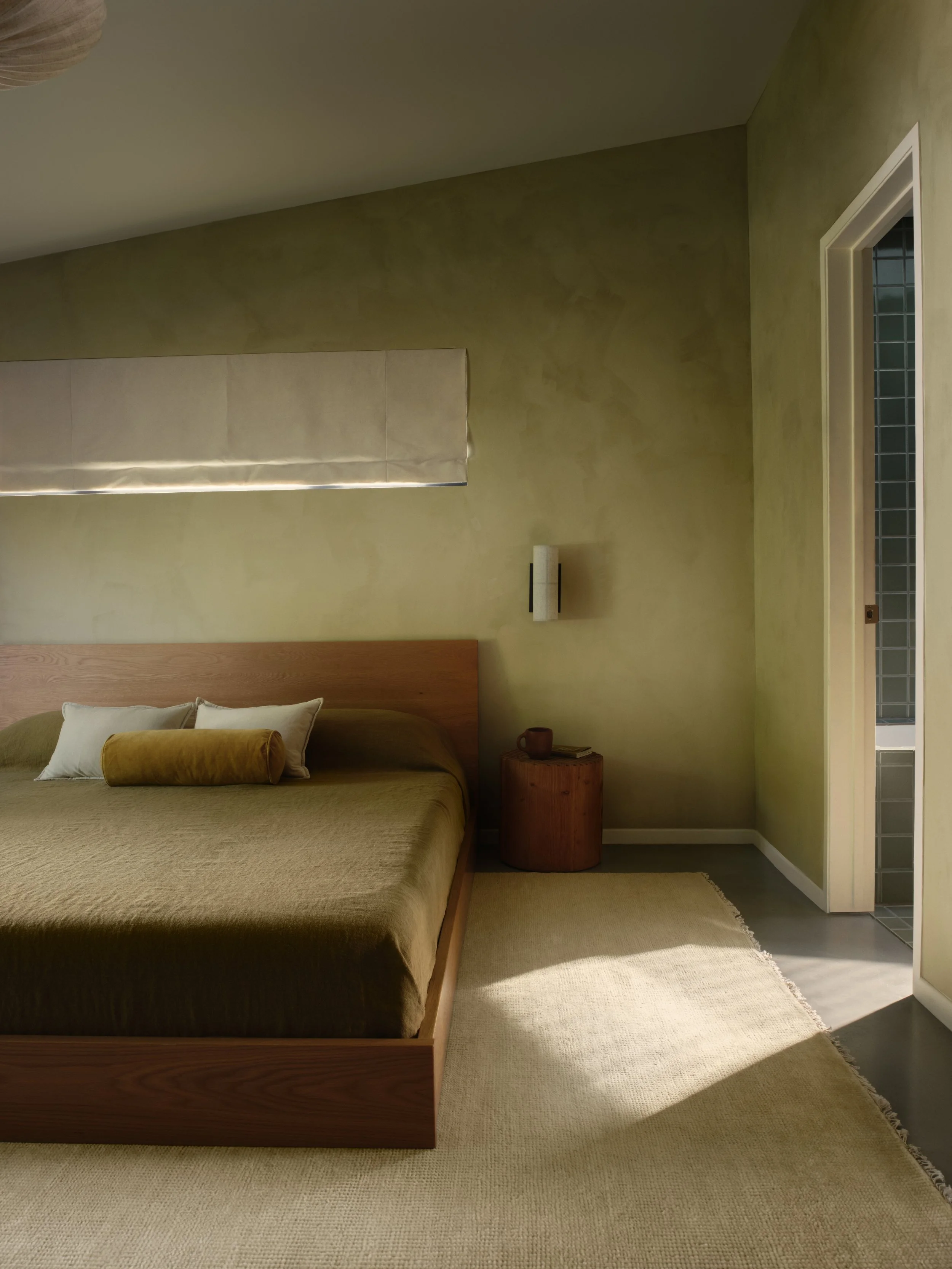 Minimalist bedroom with olive green walls, a wooden platform bed, earthy bedding, and a small side table with a mug.