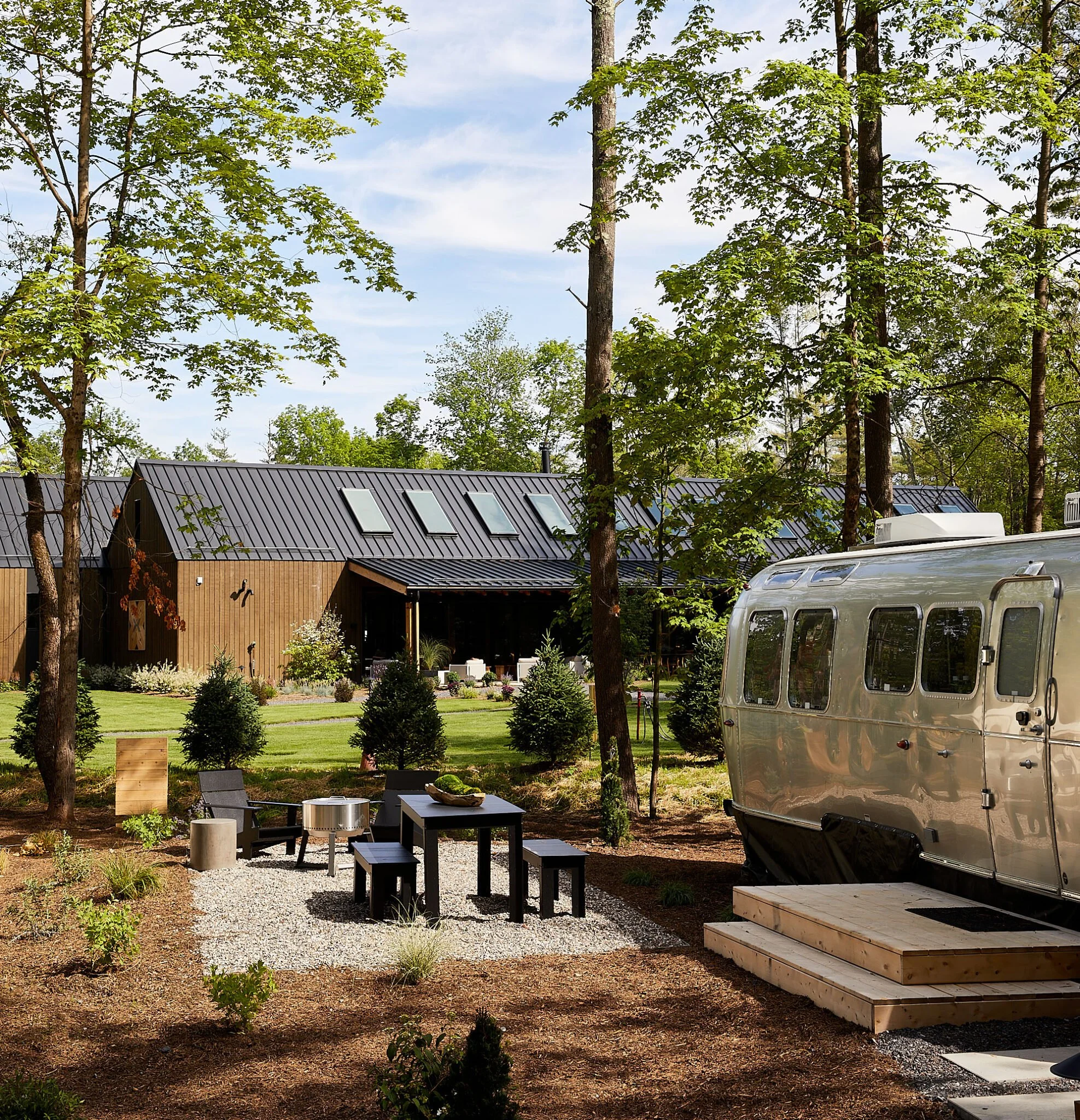 Modern cabin with sloped roof next to a silver Airstream trailer in a wooded setting, featuring outdoor seating and a fire pit on a gravel area.