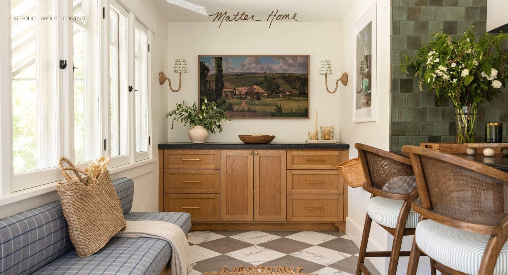 A cozy interior space featuring wooden cabinets with a countertop, a landscape painting on the wall, and two wall sconces. There is a blue cushioned bench with a woven basket on it near windows to the left. On the right, there is a small bar area with wooden chairs, green tiled wall, and a vase with flowers.