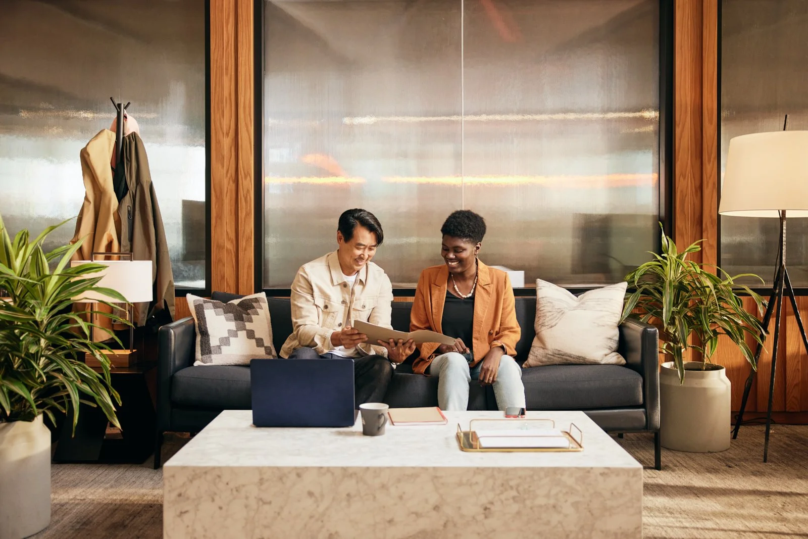 Two people sitting on a couch in an office, looking at documents, with plants and a laptop on a table in front of them.