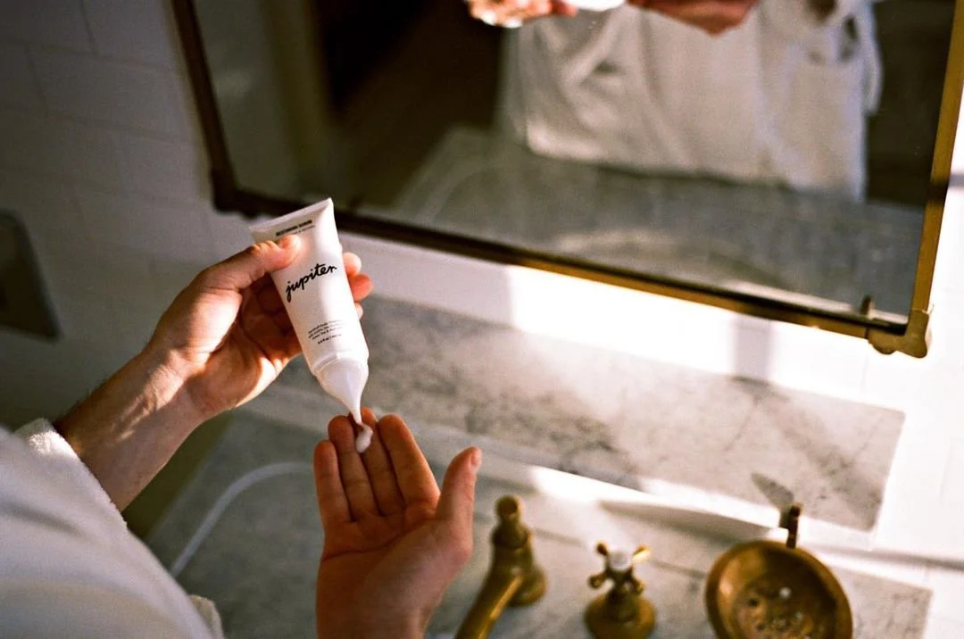 Person squeezing cream onto hand from a tube labeled 'Jupiter' in a bathroom with brass fixtures and a marble countertop.
