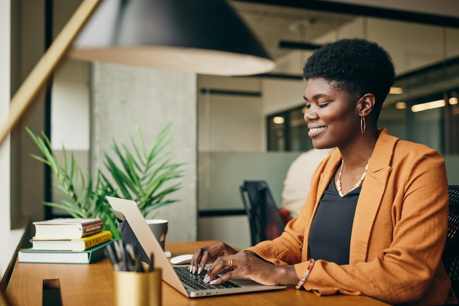 Woman in orange blazer working on a laptop at a desk in an office setting, with books, a plant, and a cup nearby.