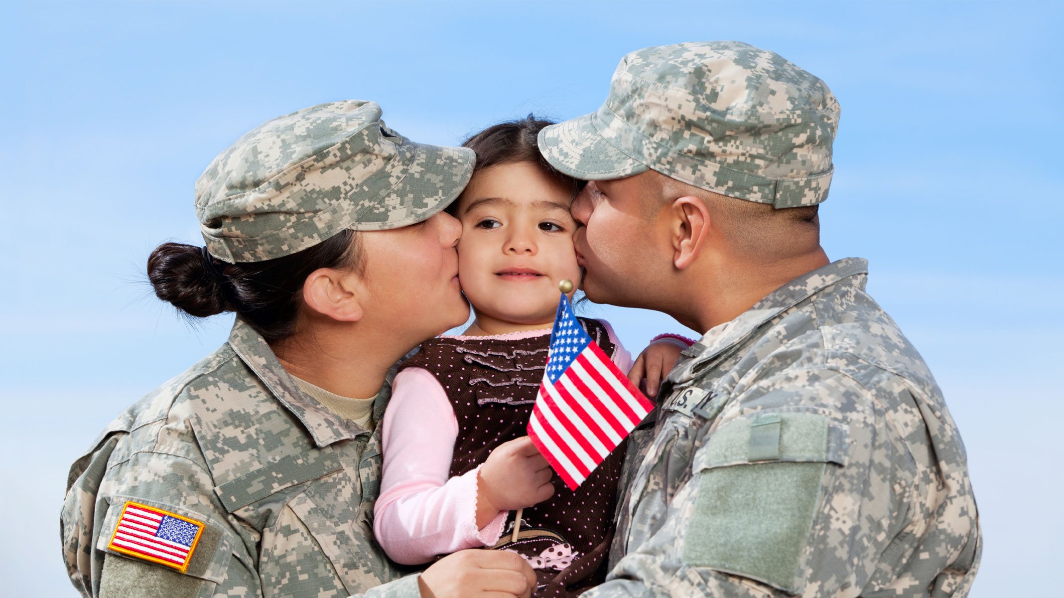 Two soldiers wearing camouflage uniforms kiss a girl holding an American flag against a blue sky.
