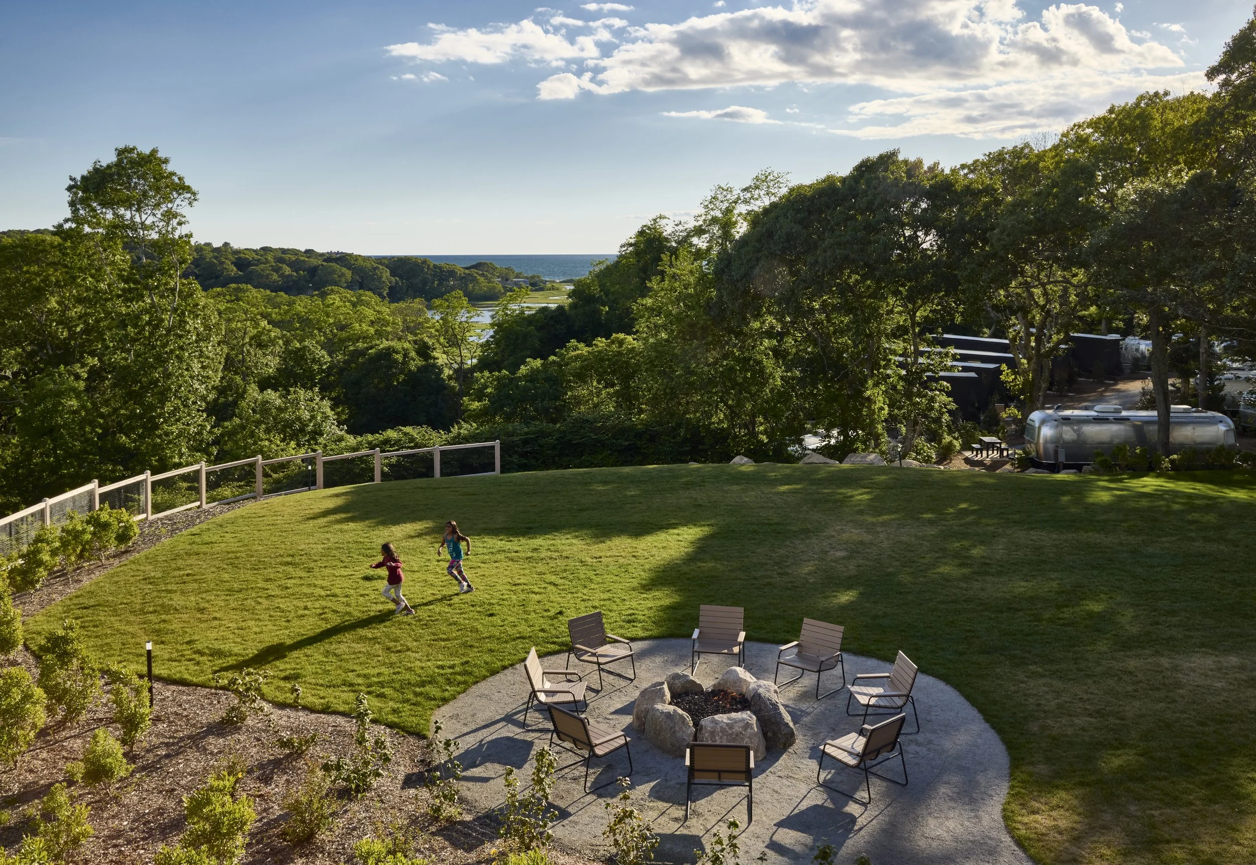 A large grassy backyard with a circular fire pit surrounded by wooden chairs. Two children are running on the grass. Trees and an ocean view are in the background. It's a sunny day with a few clouds in the sky.