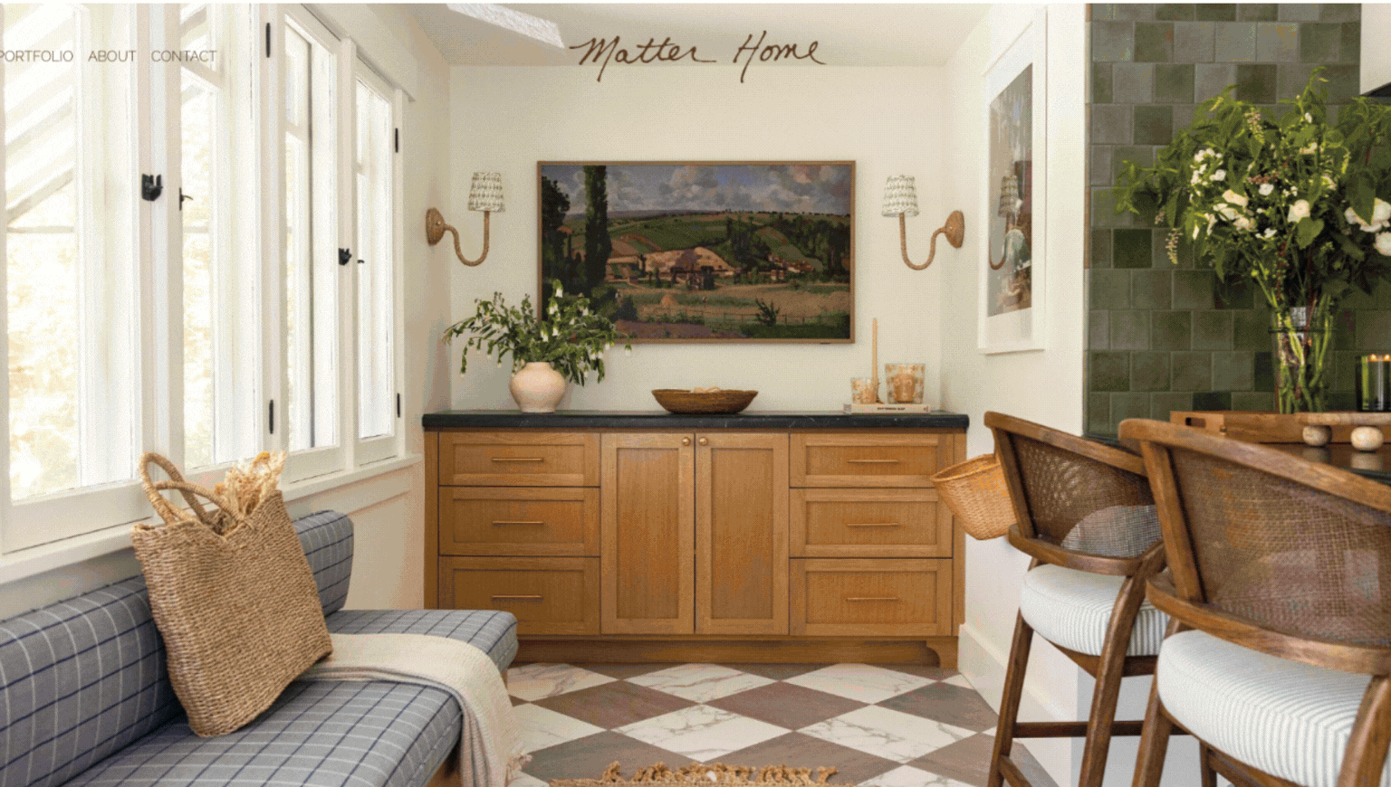 Interior view of a cozy, modern kitchen with wooden cabinetry, a checkered tile floor, a vase of flowers, a basket, framed artwork, and natural light from large windows.
