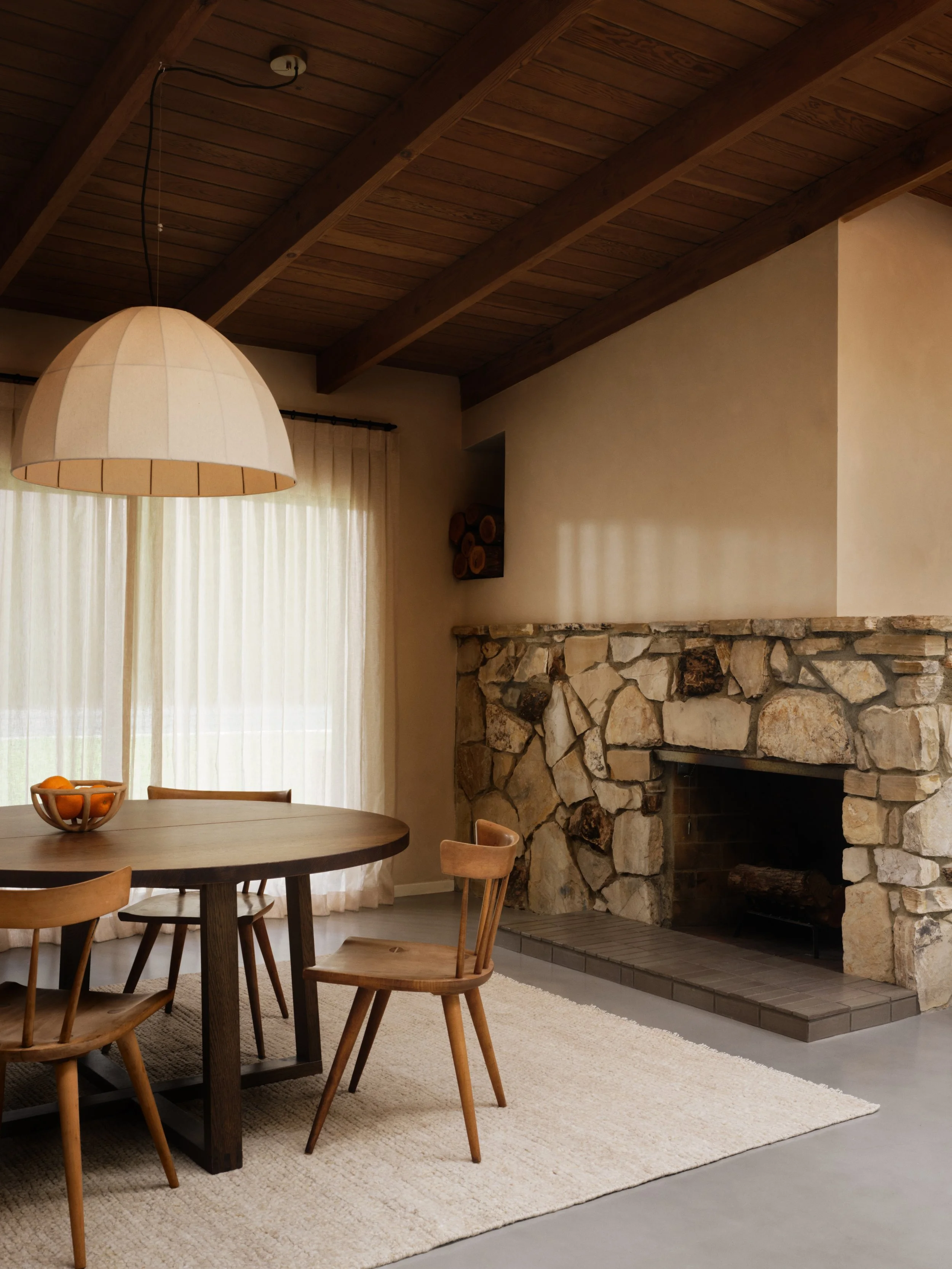 Cozy interior with a wooden round dining table, four wooden chairs, a large pendant lamp, stone fireplace, log storage, and sheer curtains.