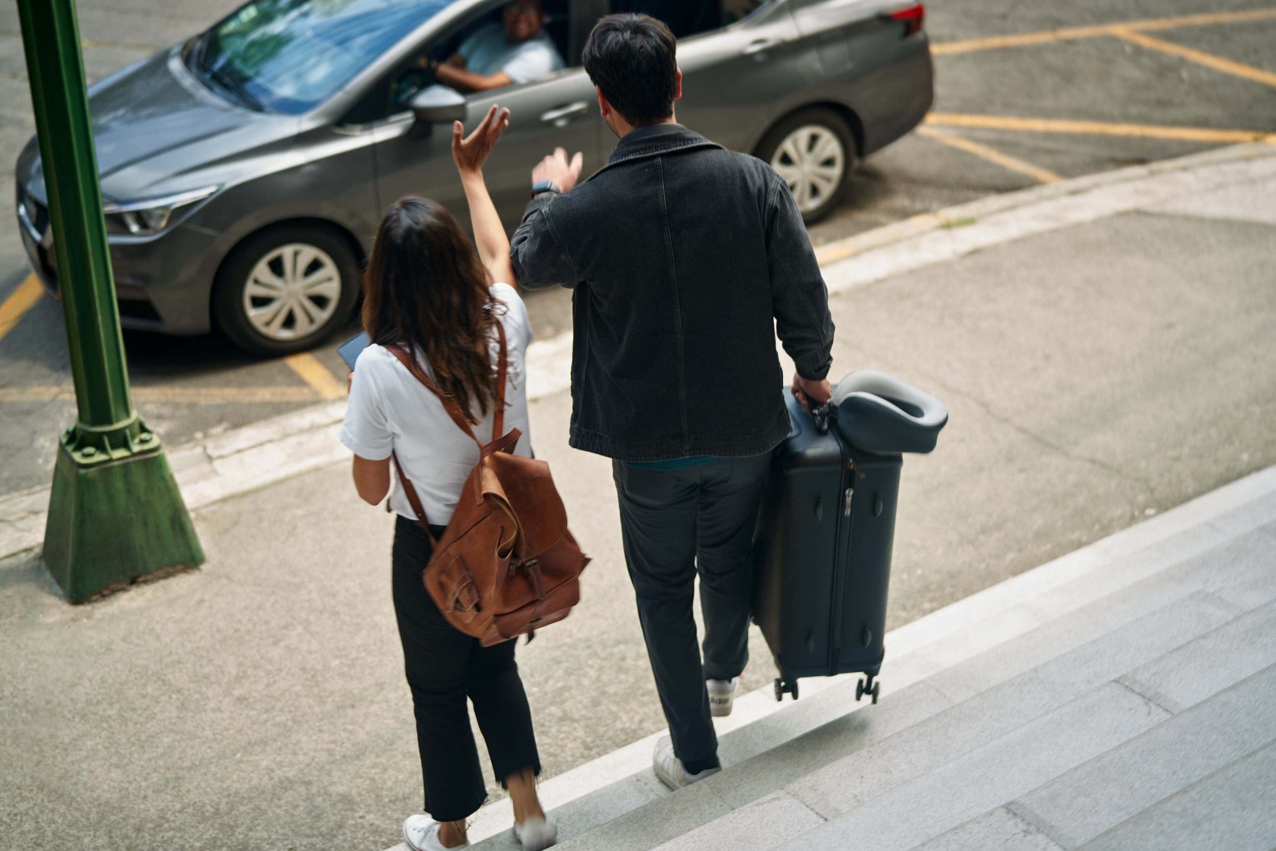 Two people with luggage waving at a driver in a parked car as they walk on a sidewalk.