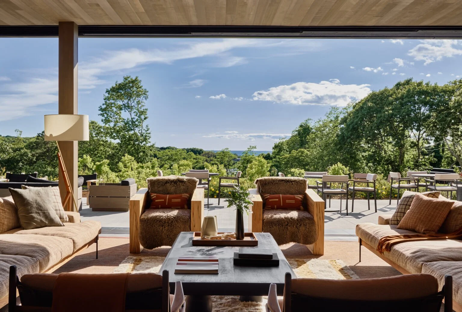 A luxurious living room with comfortable sofas and armchairs facing a large open terrace with outdoor seating, overlooking a scenic view of trees and blue sky, under a wooden ceiling.