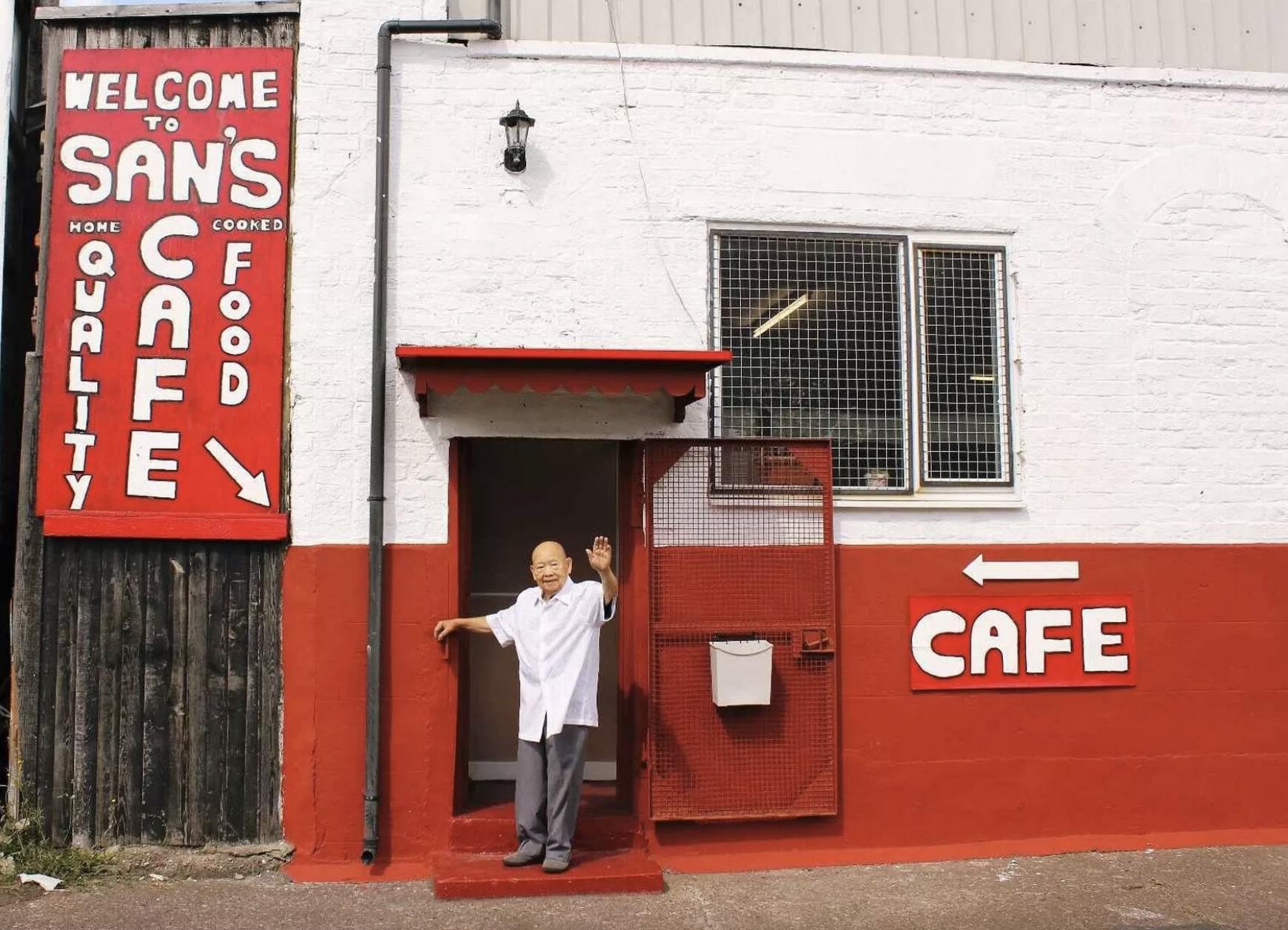 A man in a white coat standing at the entrance of a cafe, waving. The building has white and red walls with signs that read 'Welcome to San's' and 'Cafe,' with an arrow pointing left towards the cafe entrance.