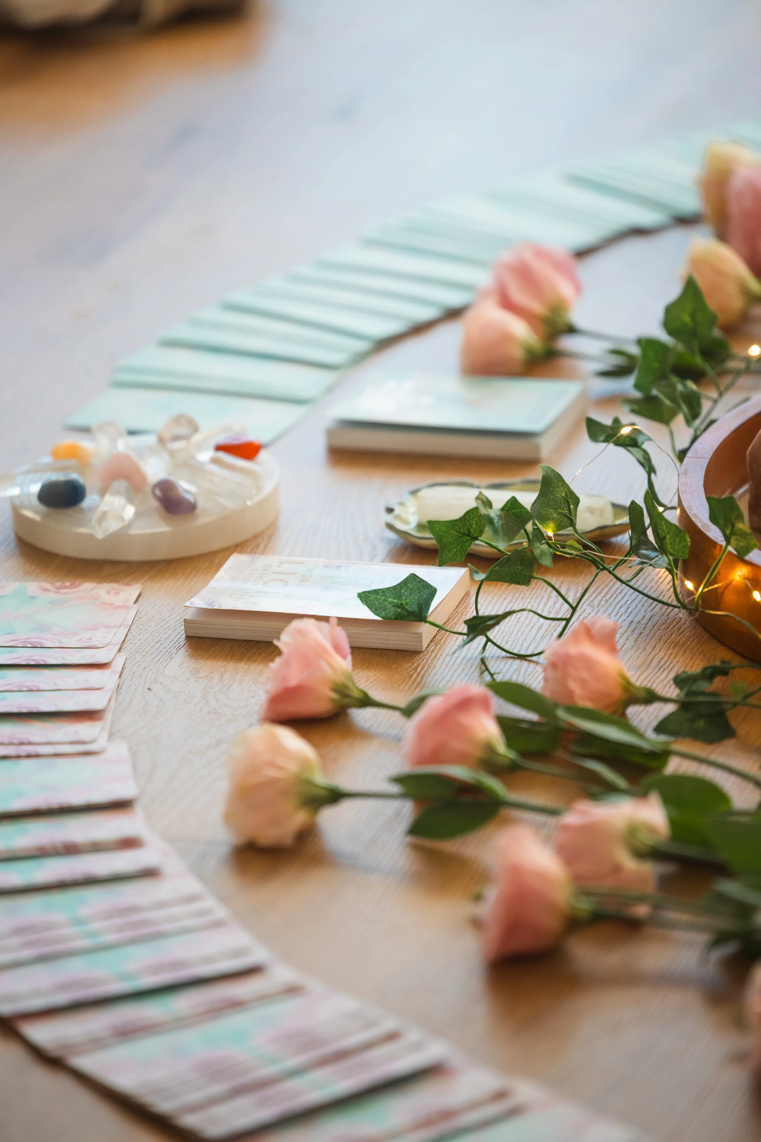 Floral arrangement with pink roses, fake green leaves, fairy lights, a stack of small cards, a round tray of beads, and some open cards with pastel-colored designs on a wooden table.