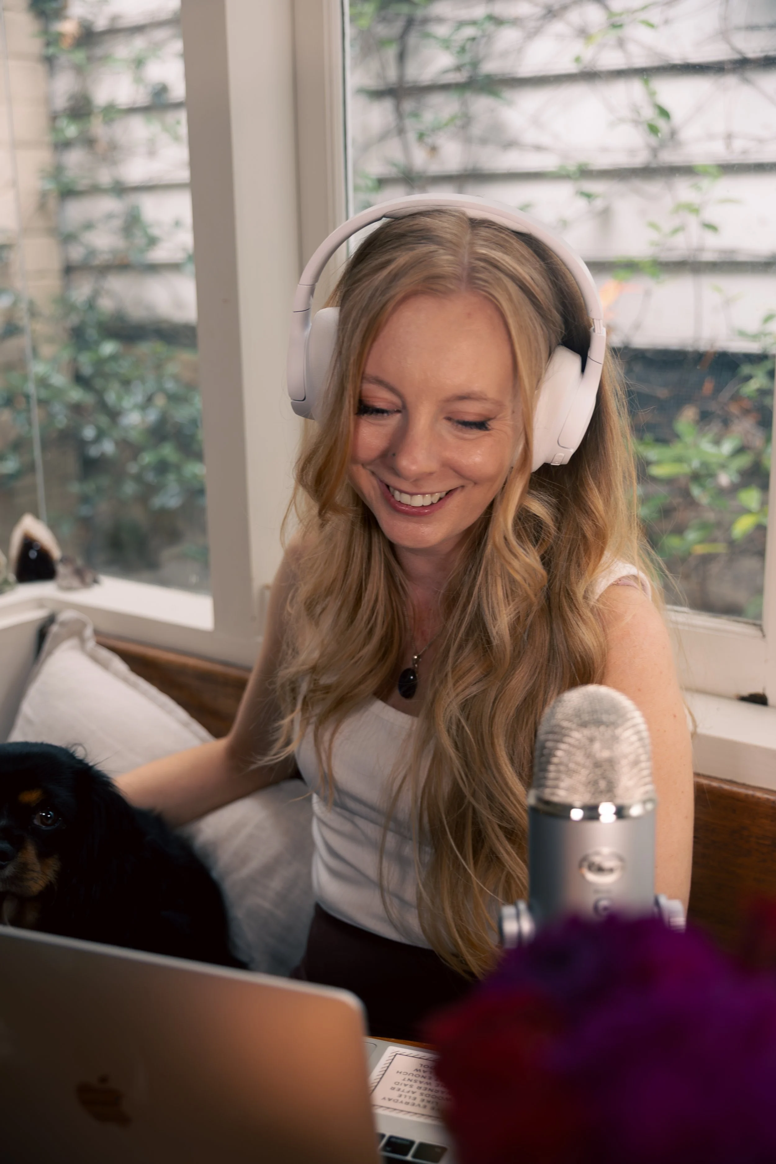 A woman with long blonde hair wearing white headphones smiling, sitting at a desk with a laptop, a microphone in front of her, a small dog lying on a blanket, and a window with greenery outside.
