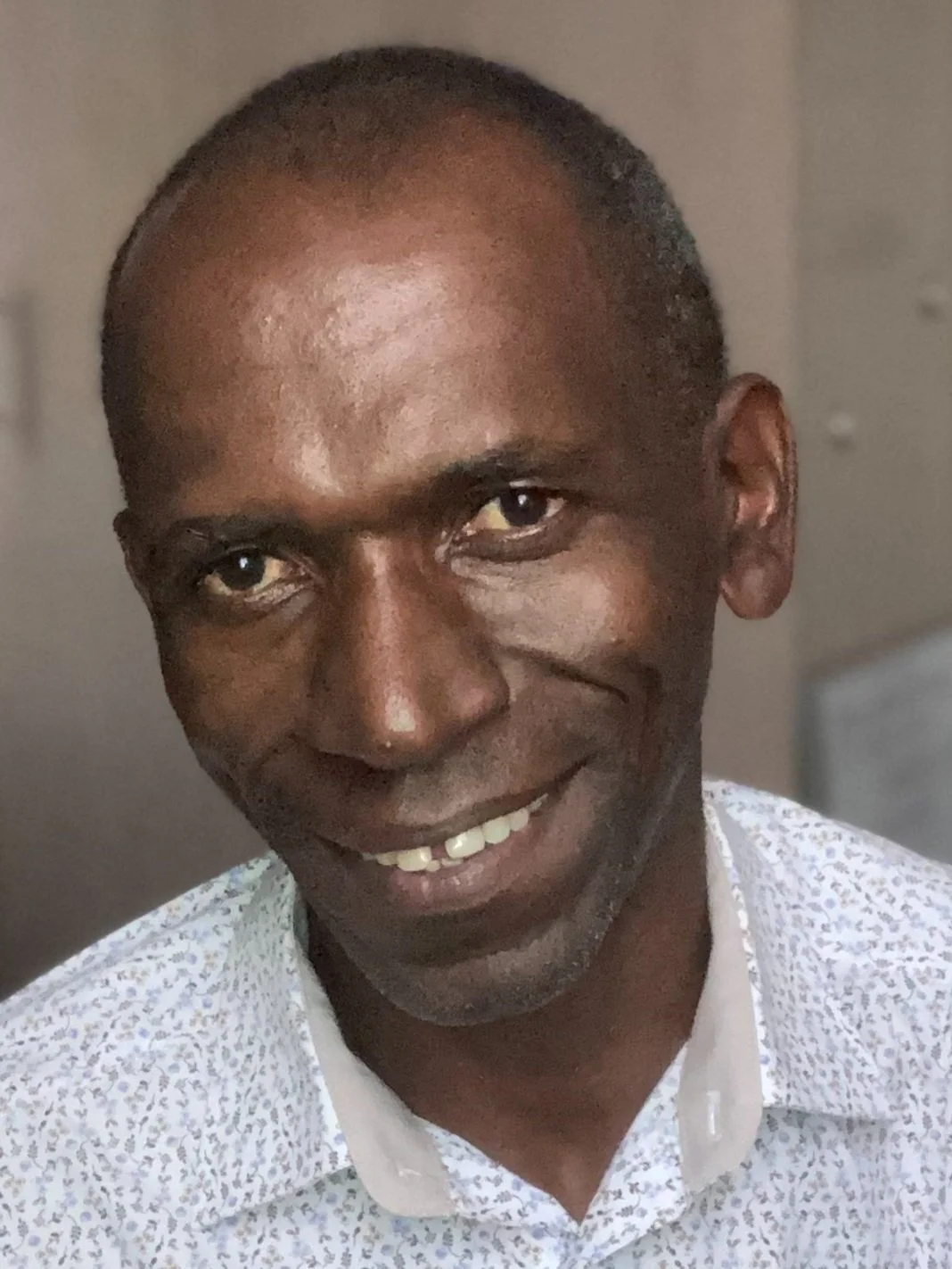 Close-up of a smiling man with dark skin, bald head, and wearing a white shirt with a subtle pattern.