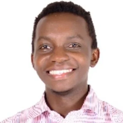 A young man with short curly hair, smiling, wearing a pink and white striped collared shirt.