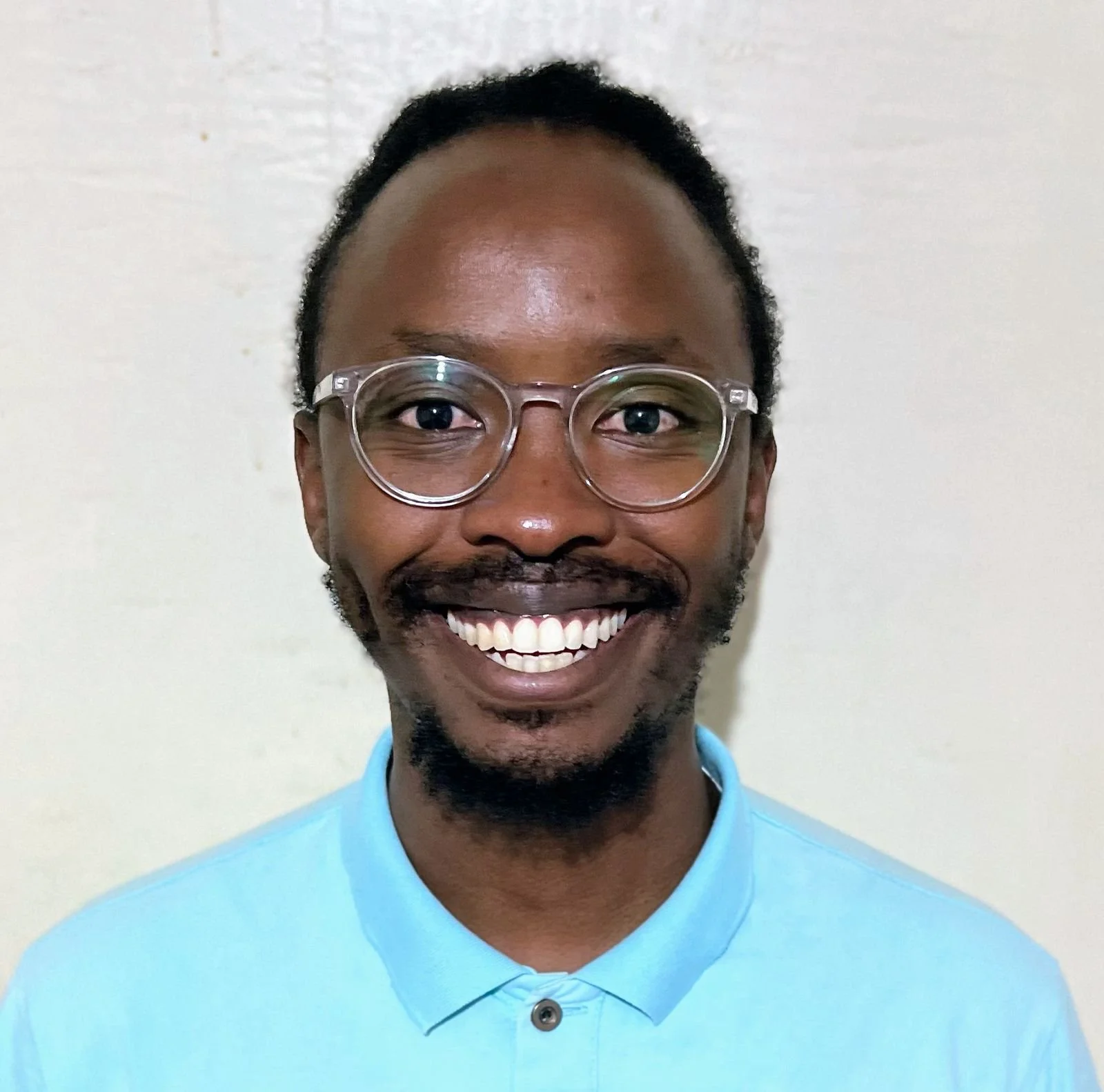 A smiling man with glasses and a beard, wearing a light blue collared shirt, standing in front of a plain off-white wall.