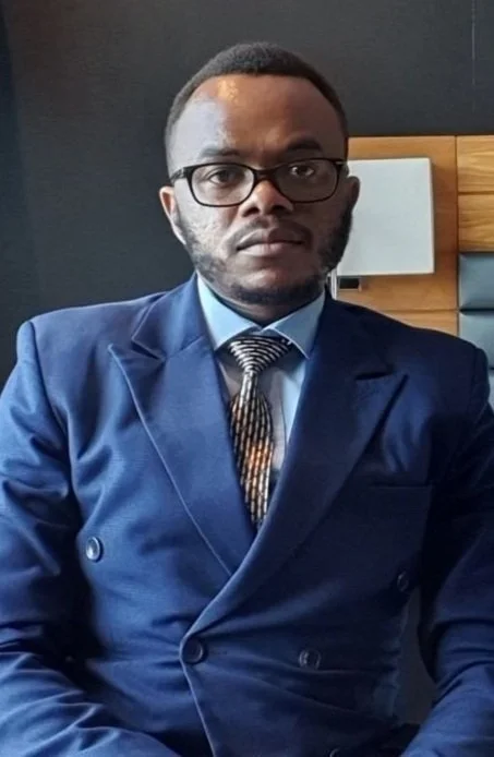 A man in a dark blue suit, light blue shirt, and striped tie, wearing glasses, sitting indoors with a serious expression.
