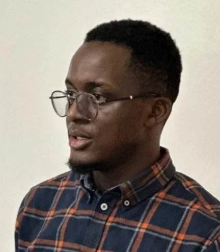 A young man wearing glasses and a plaid shirt, looking to the side, with a neutral background.