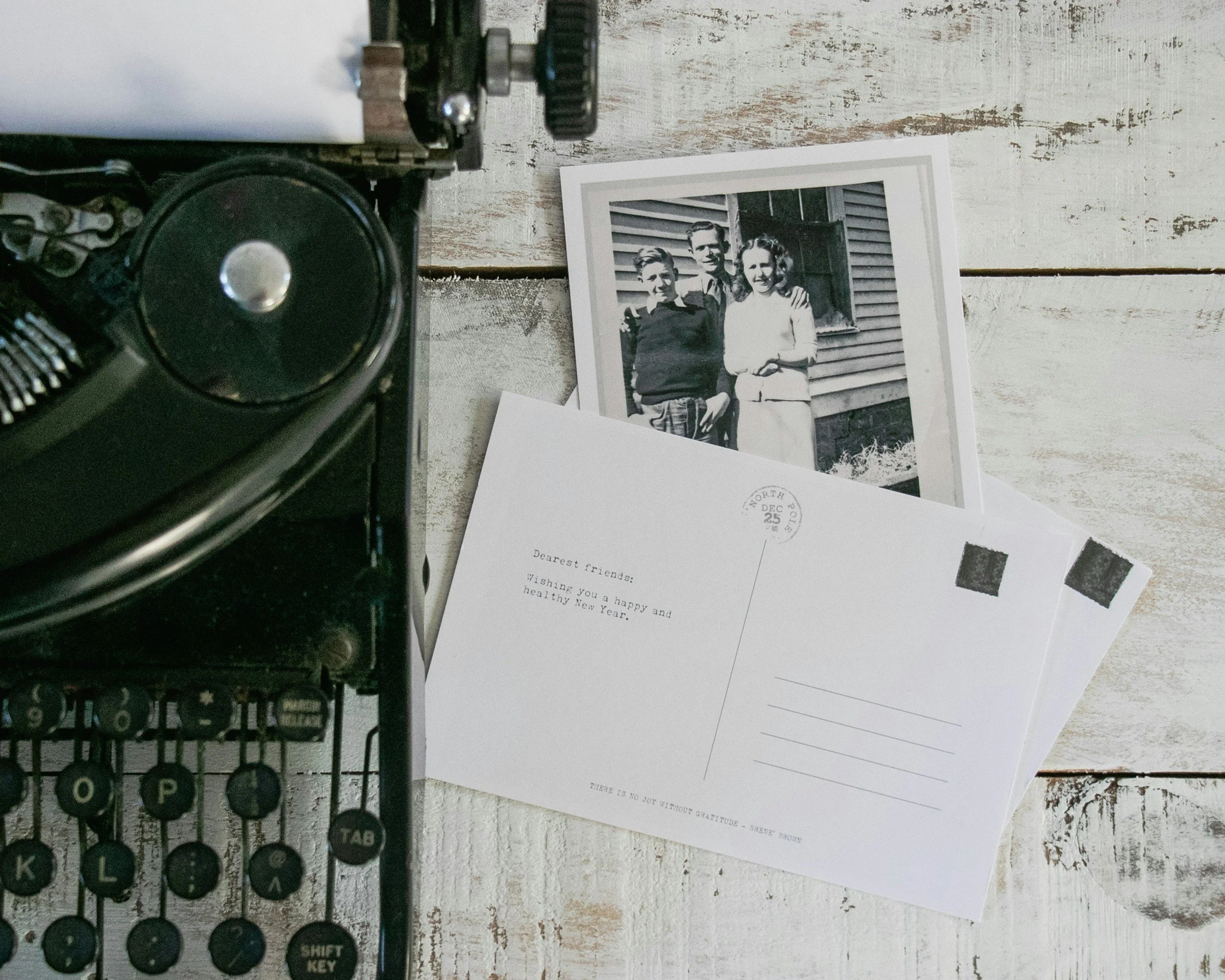 A vintage black typewriter on a distressed white wooden surface, with black and white photographs of three people and some blank postcards laid out.