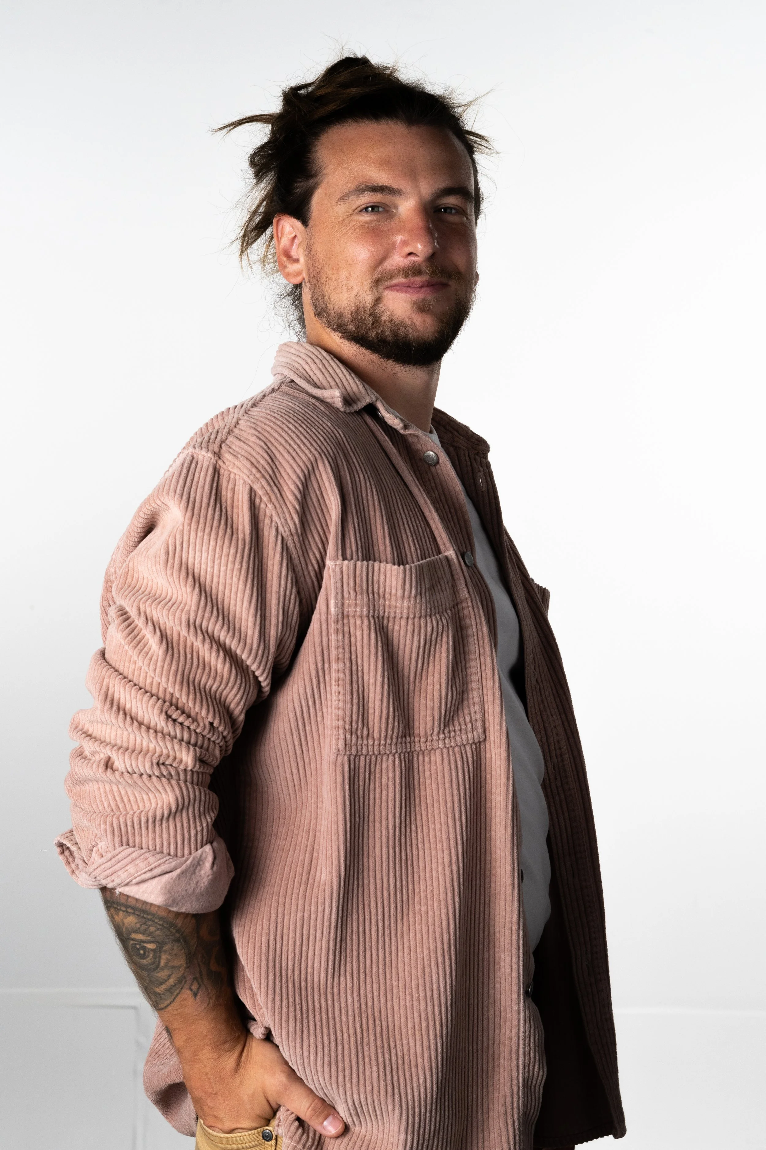 A man with a beard and tophair styled back, wearing a brown corduroy jacket over a white shirt, standing against a plain white background.
